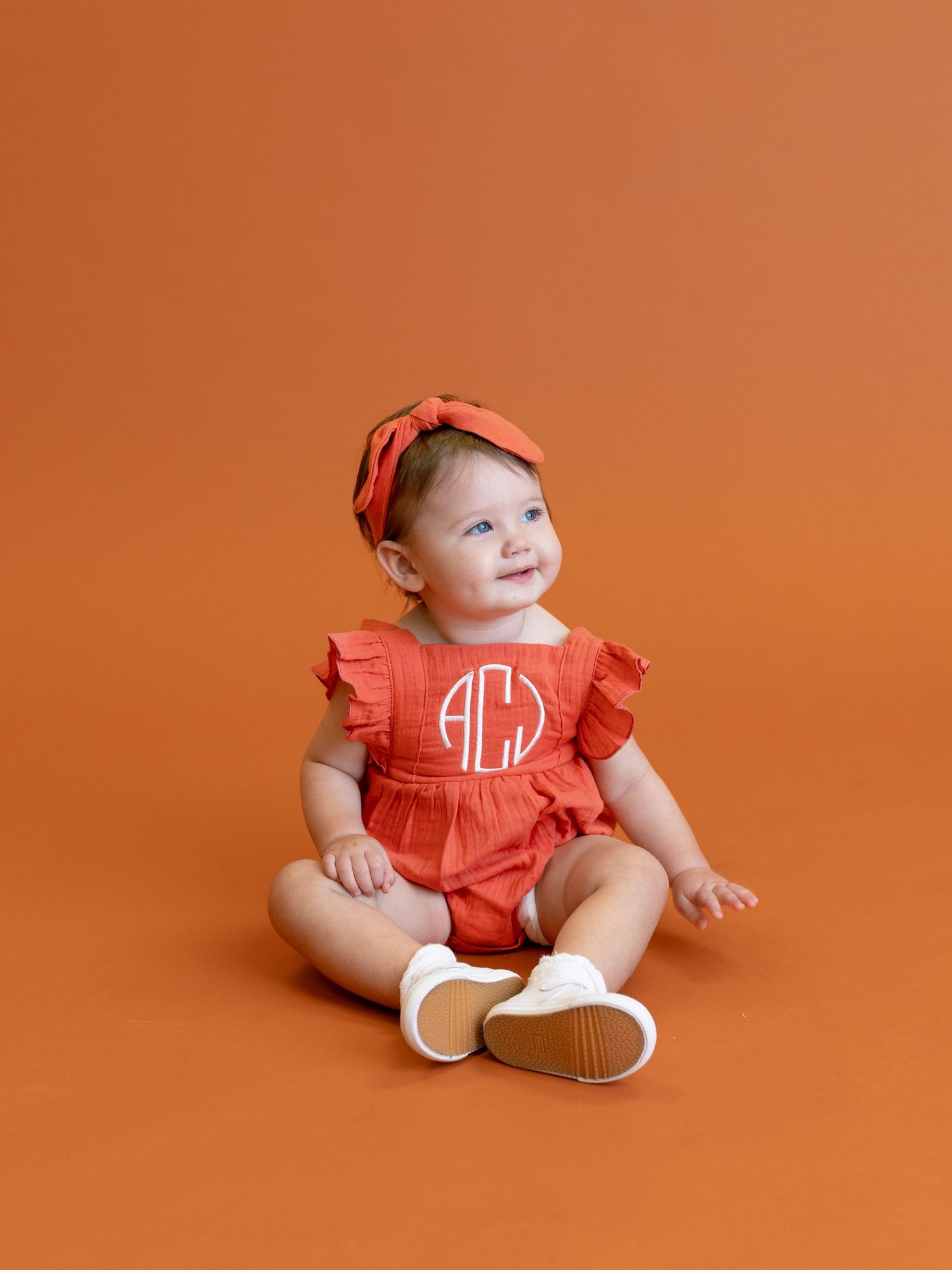Baby wearing red monogrammed outfit with ruffles and white shoes against orange background.