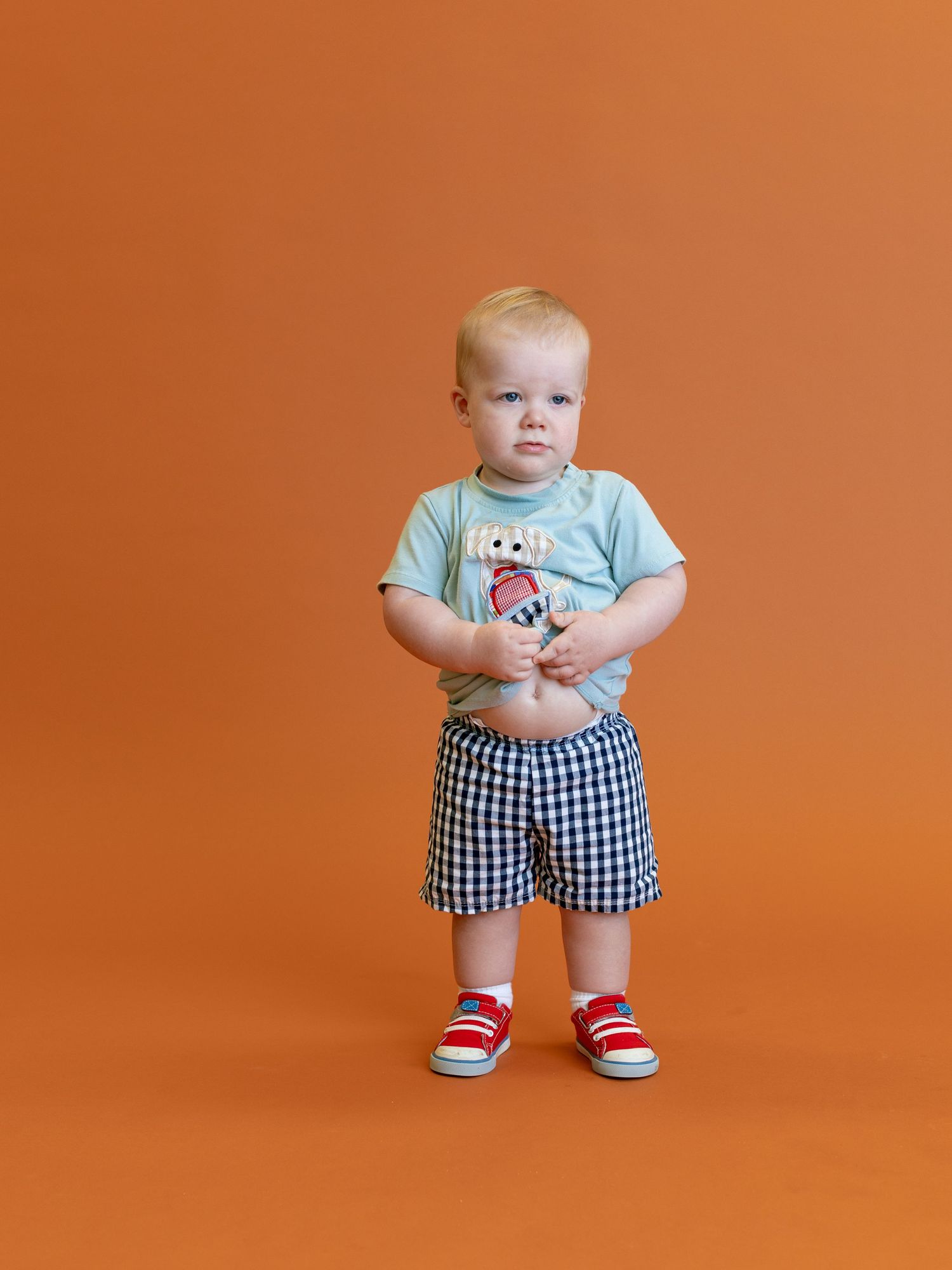 Small child in plaid shorts and graphic t-shirt standing against orange background.