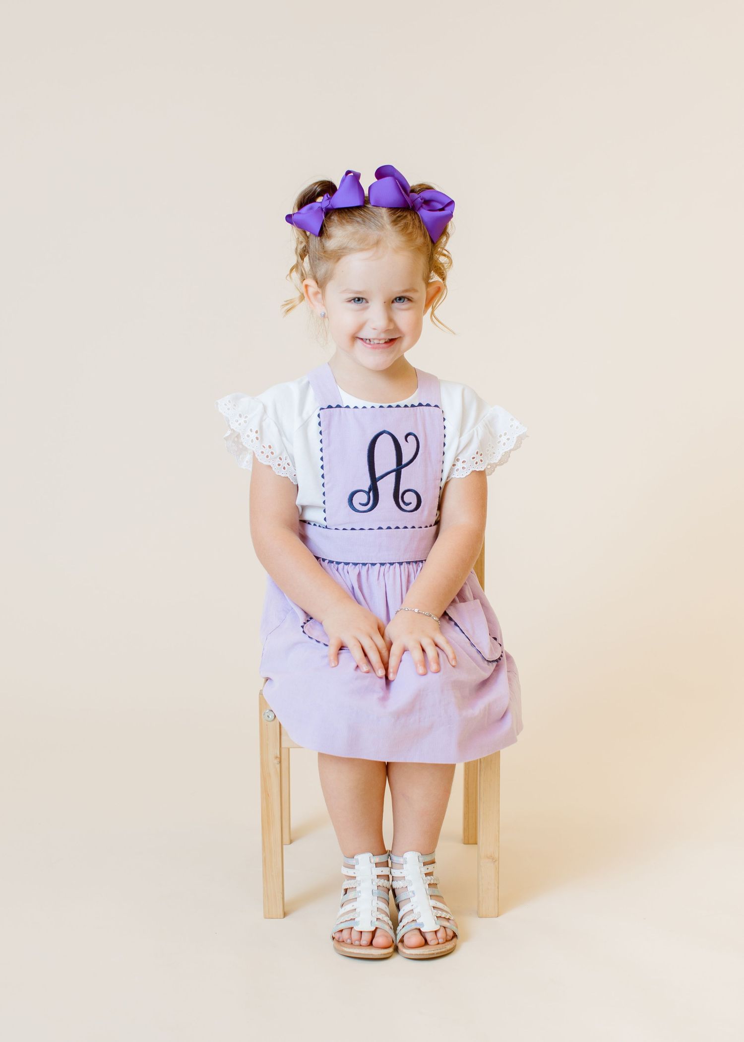Child in lavender dress with monogram and purple bow sits on wooden chair.