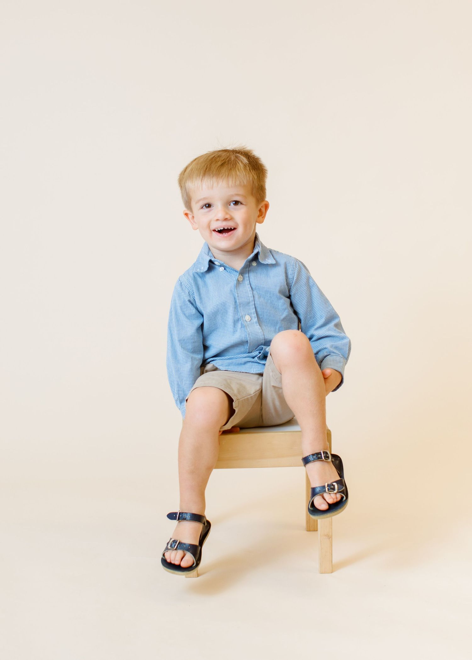 Child in blue button-down shirt sits on wooden chair wearing sandals.
