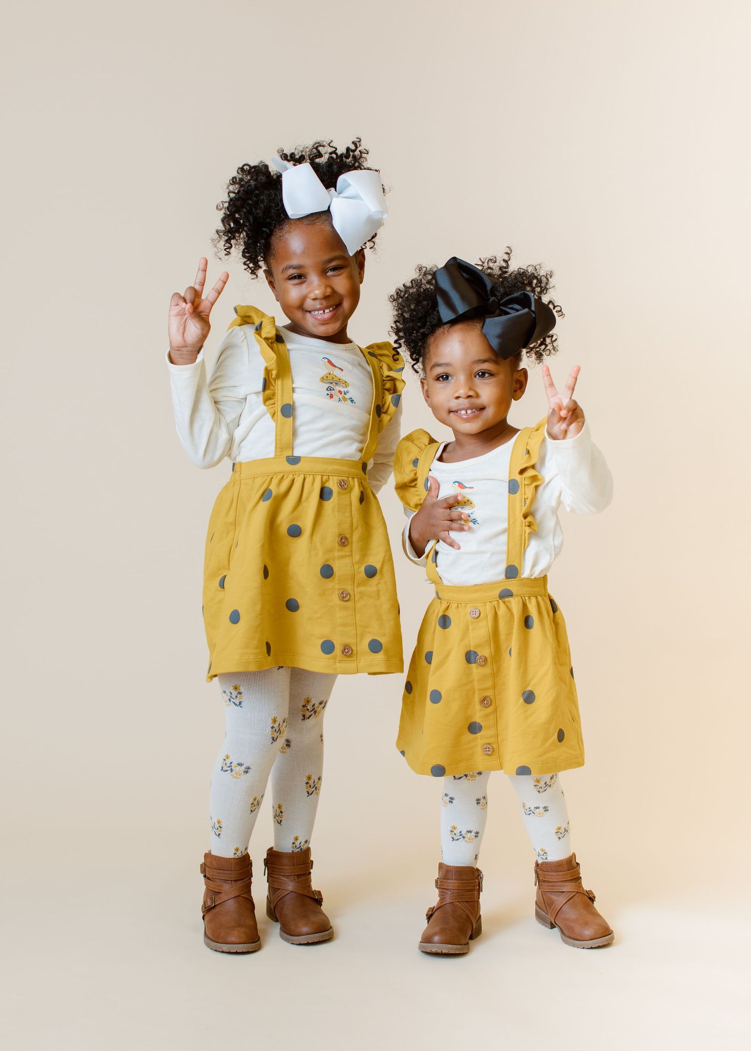 Two children in matching yellow suspender skirts and bows make peace signs while smiling.