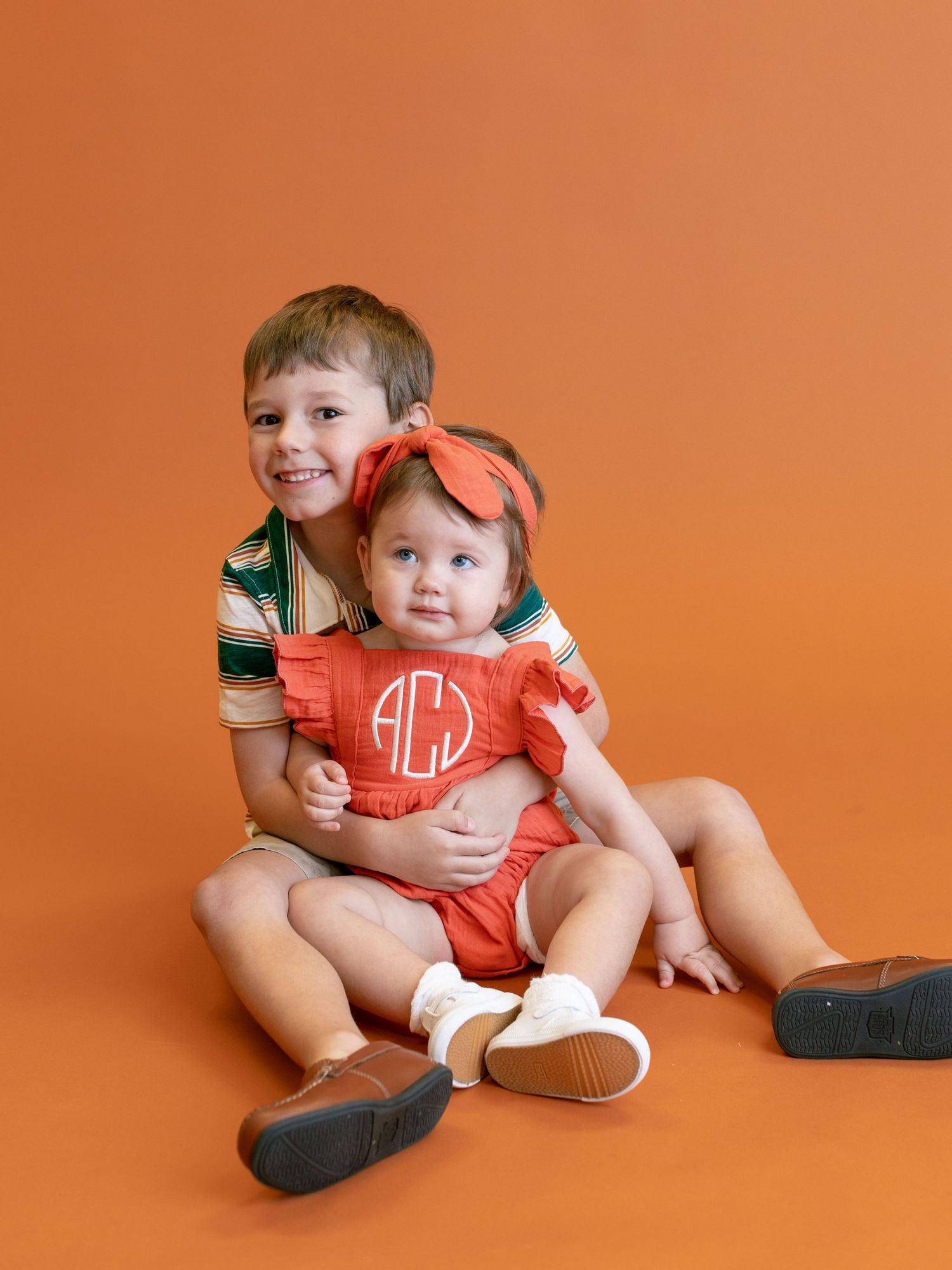 Two siblings sitting together on orange background, wearing casual clothing with monogram details.