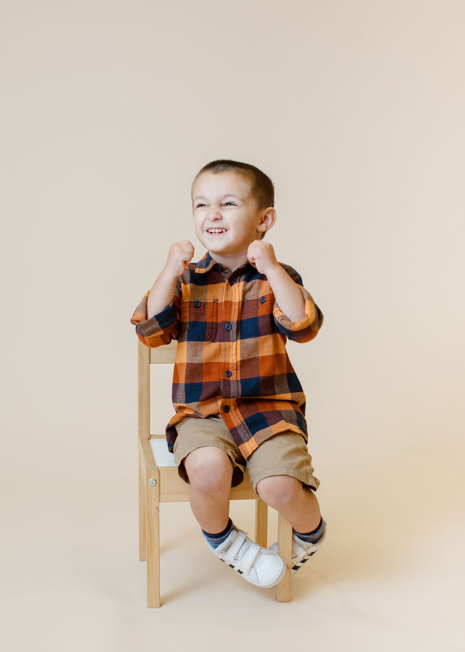 Child in orange plaid shirt and khaki shorts sitting on wooden chair against beige background.
