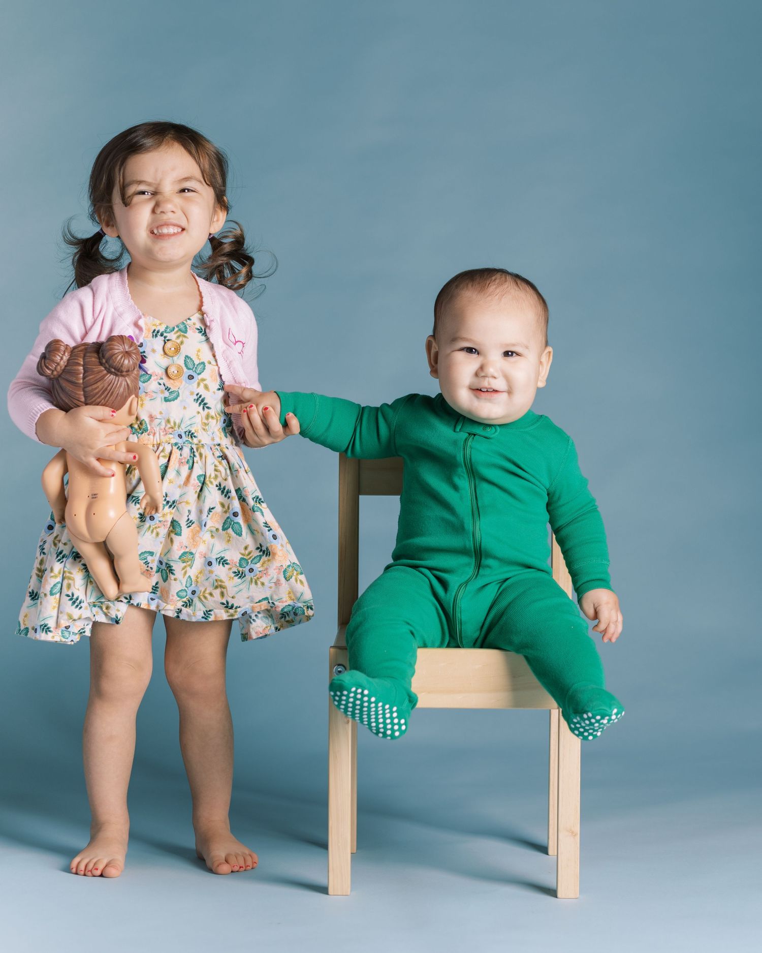 Two children posing together, one holding stuffed toy against blue background.