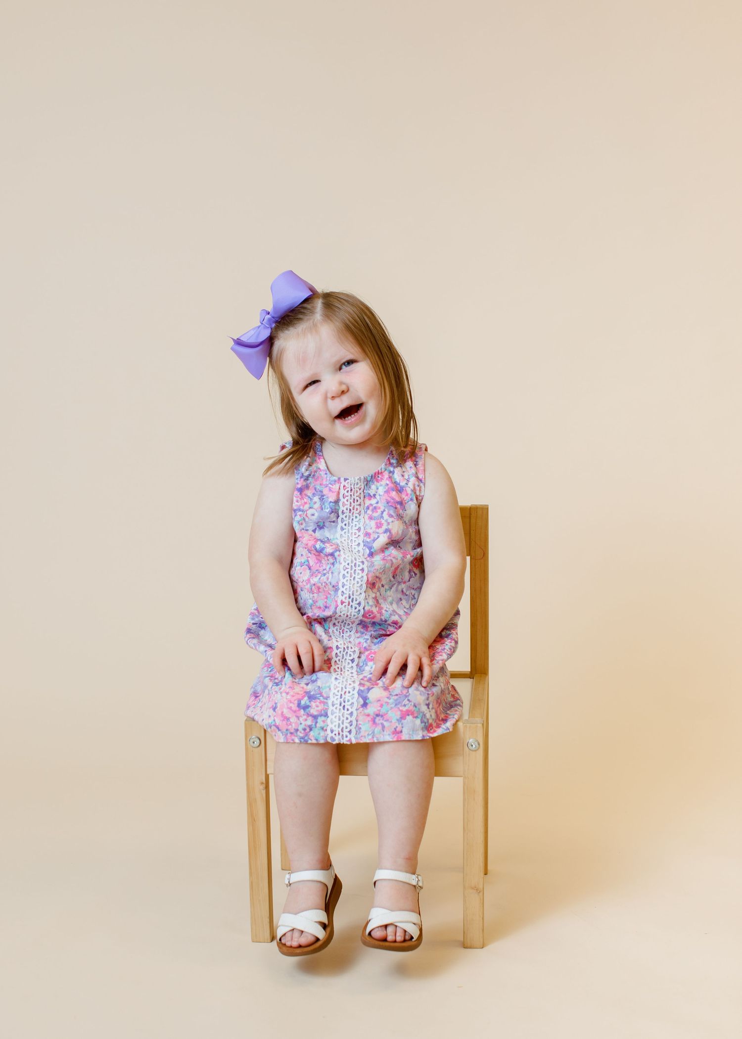 Child in floral dress and purple bow sits on wooden chair with joyful expression.
