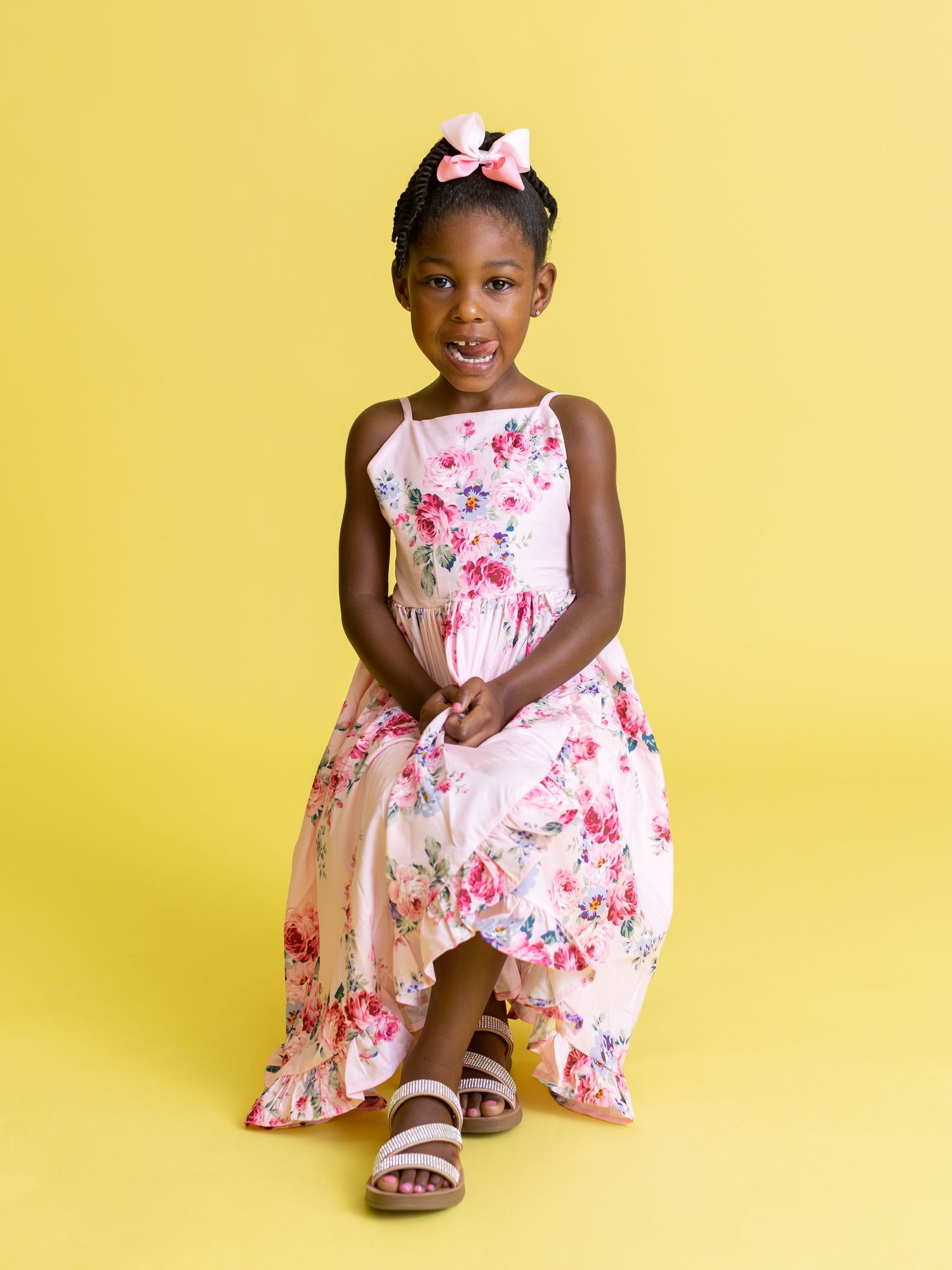 Child in floral pink dress and sandals seated against yellow background.