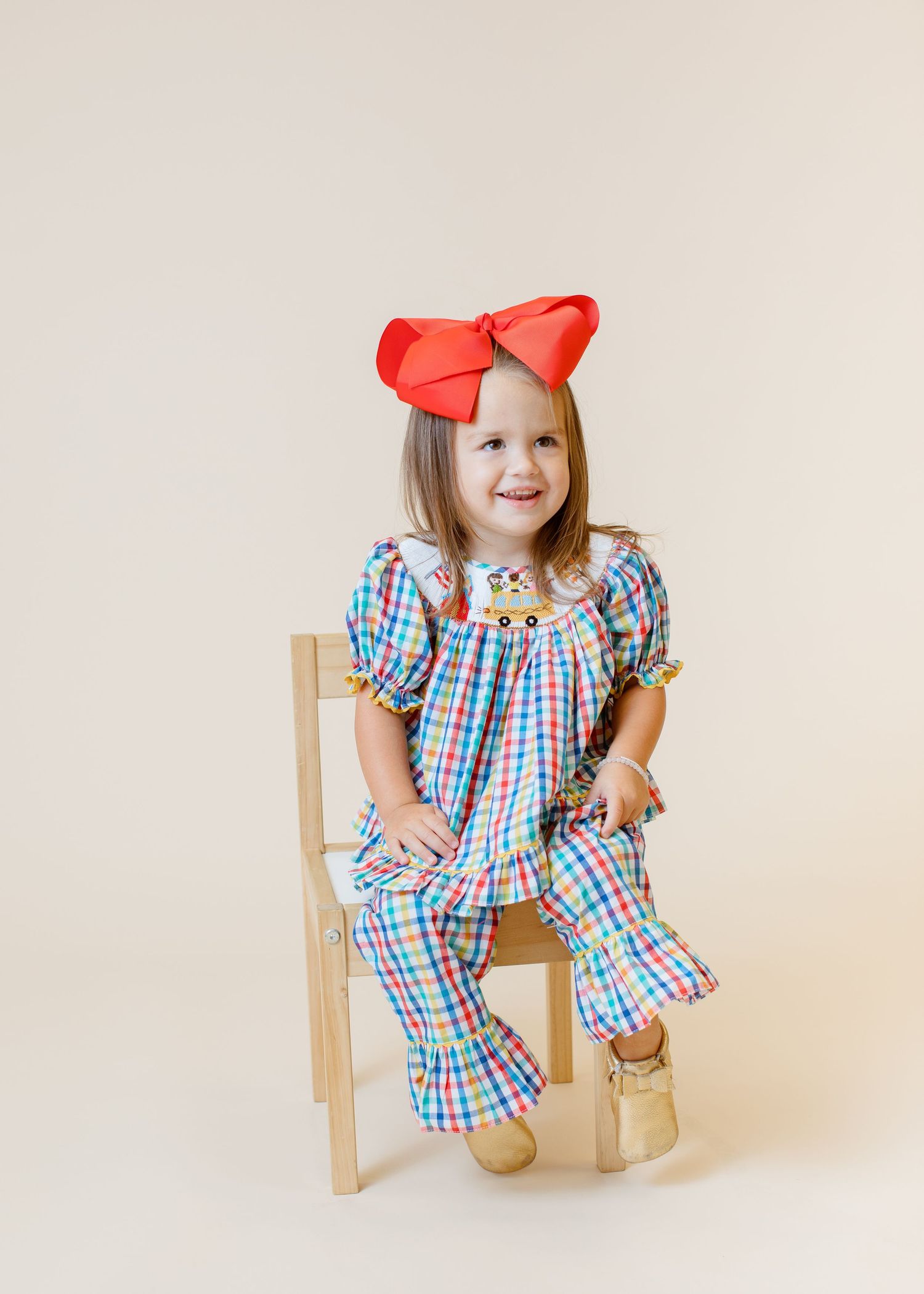 Child in colorful gingham dress and red bow sitting on wooden chair.
