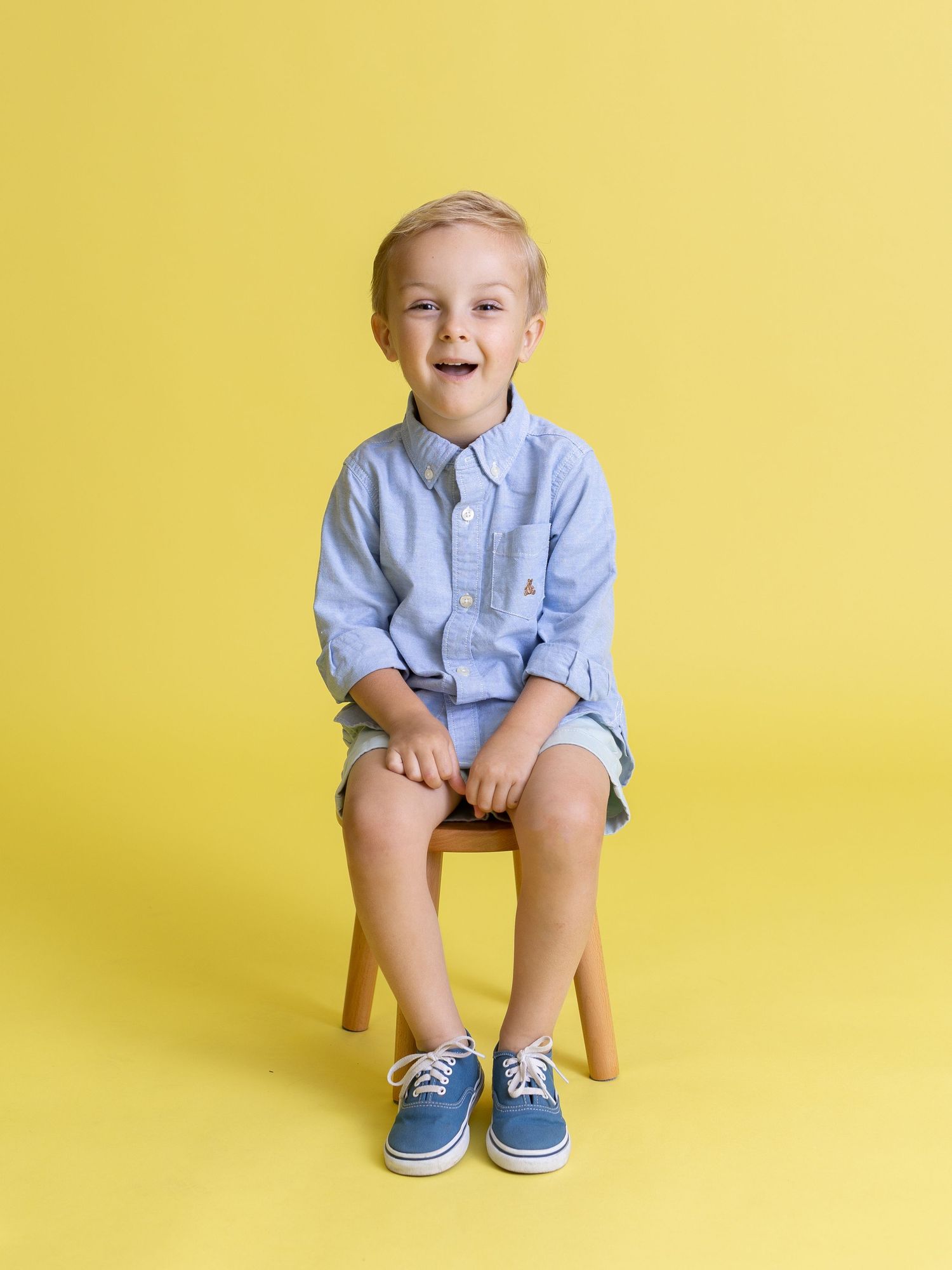 Child in light blue button-down shirt sitting on wooden stool against yellow background.