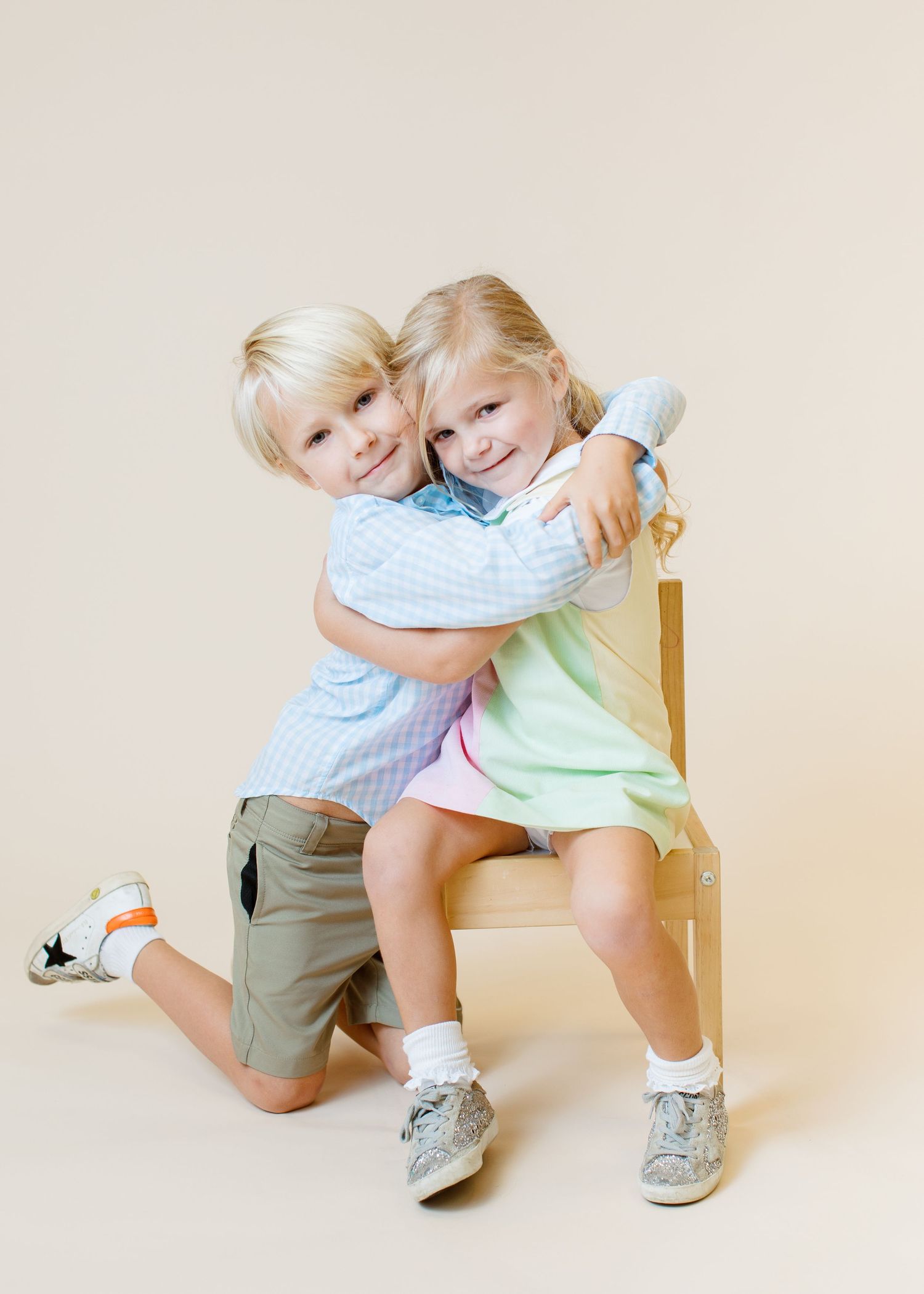 Two children hugging and smiling while sitting on wooden chair against neutral backdrop.