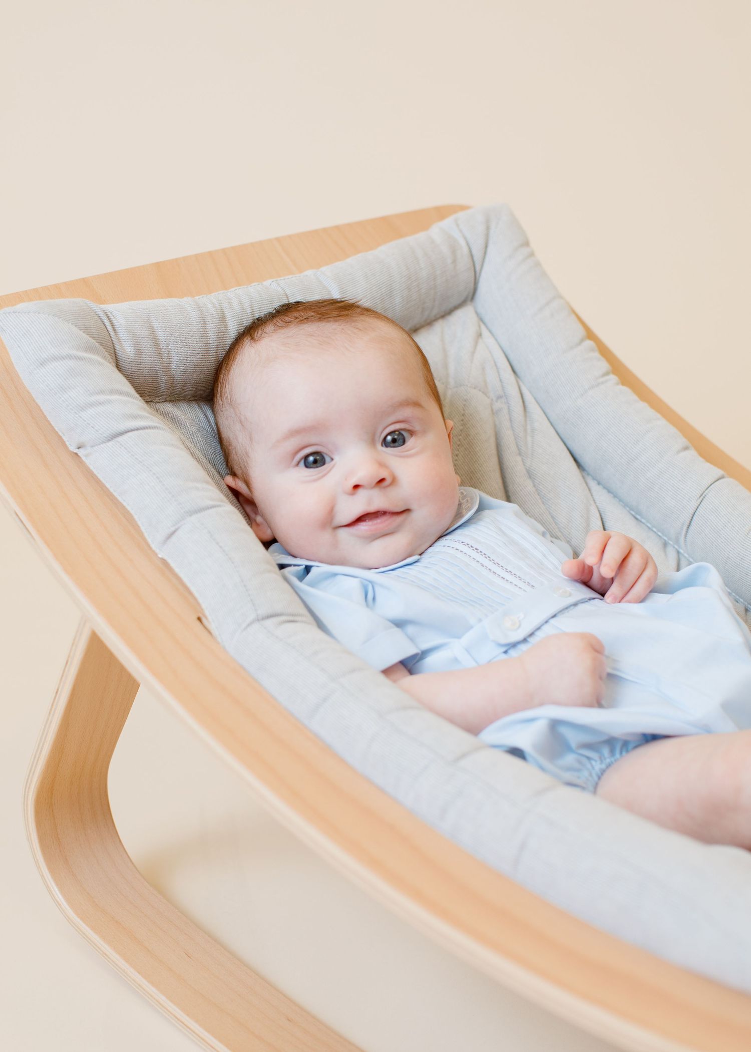 Smiling baby in light blue outfit lying comfortably in a curved wooden bouncer with grey cushioning.