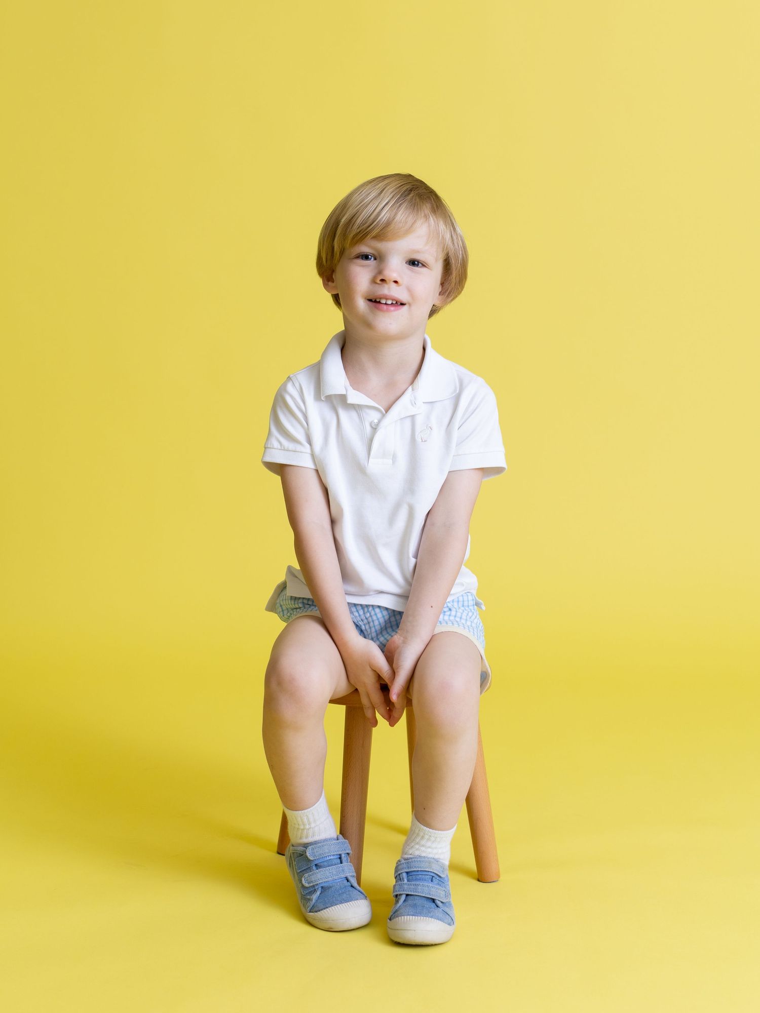 Child in white polo shirt and blue socks sitting on wooden stool against yellow background.