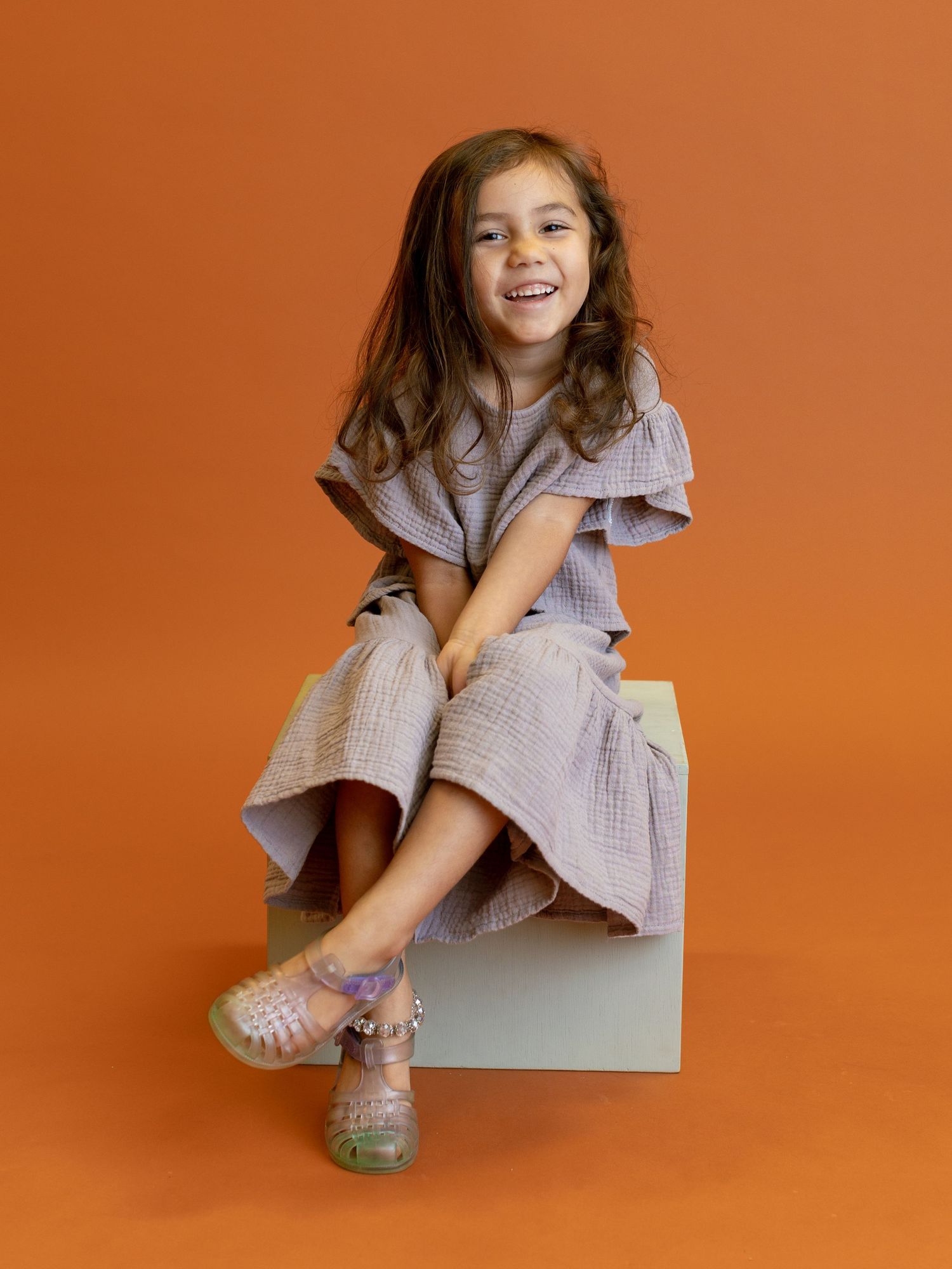 Child in lavender dress sitting on white cube against orange background.