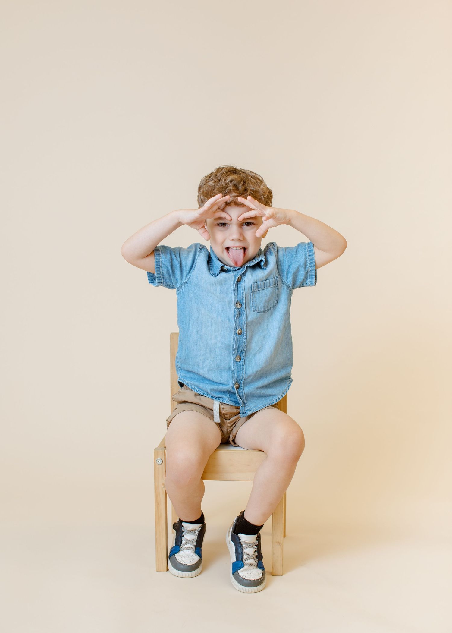 Child in denim shirt making playful gestures while sitting on wooden chair.