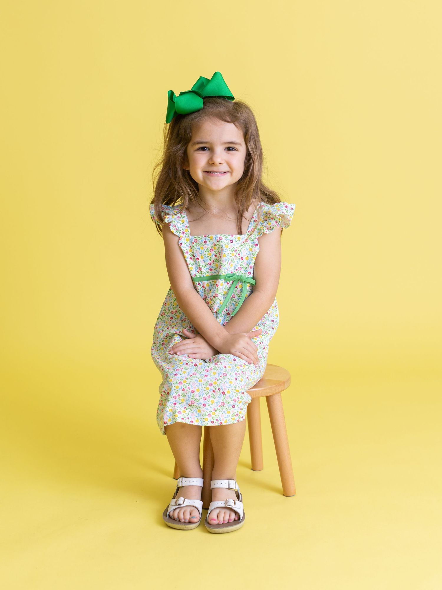 Child wearing floral dress and green crown sitting on wooden stool against yellow backdrop.