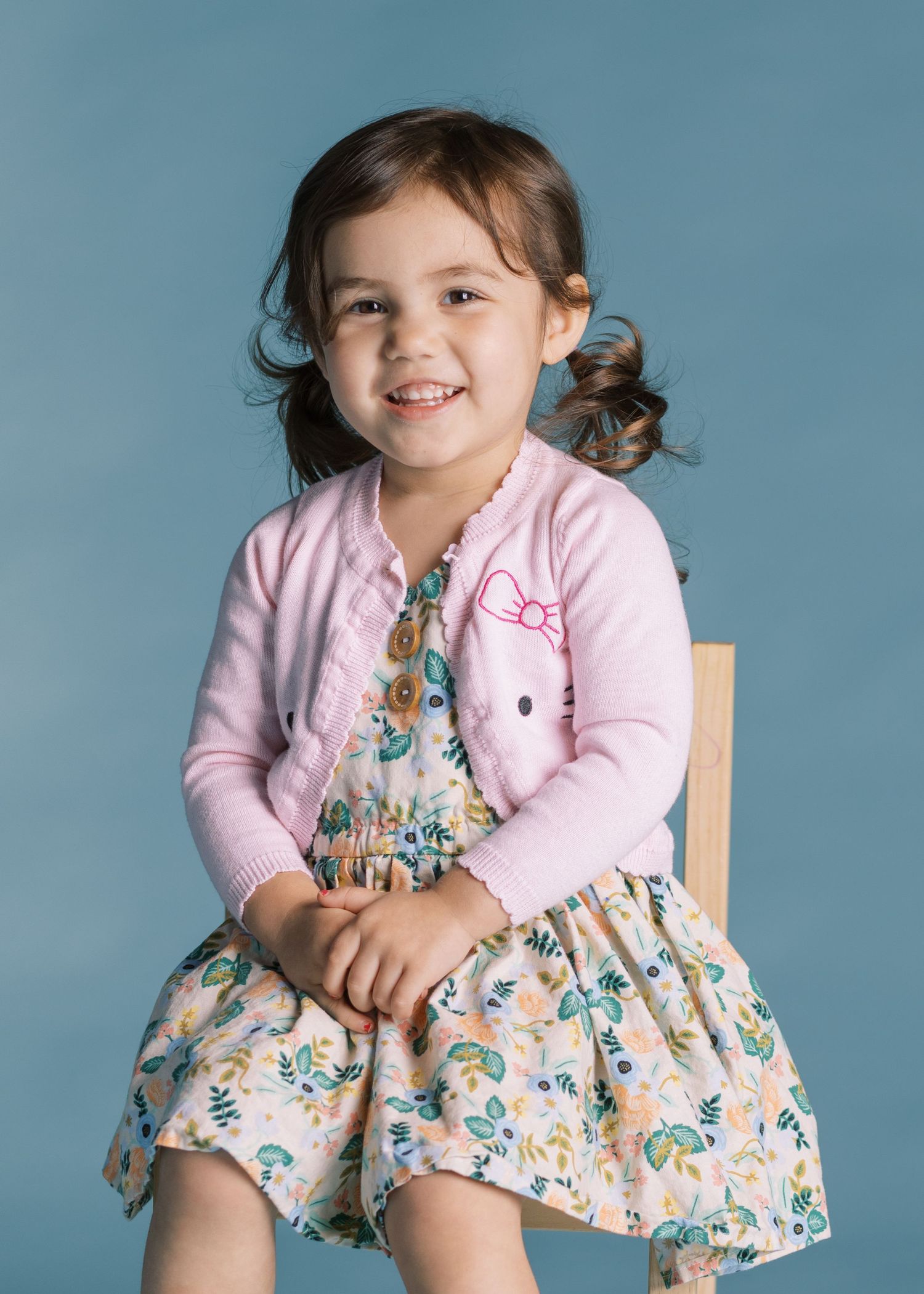 Child in floral dress and pink cardigan sitting on wooden chair against blue background.