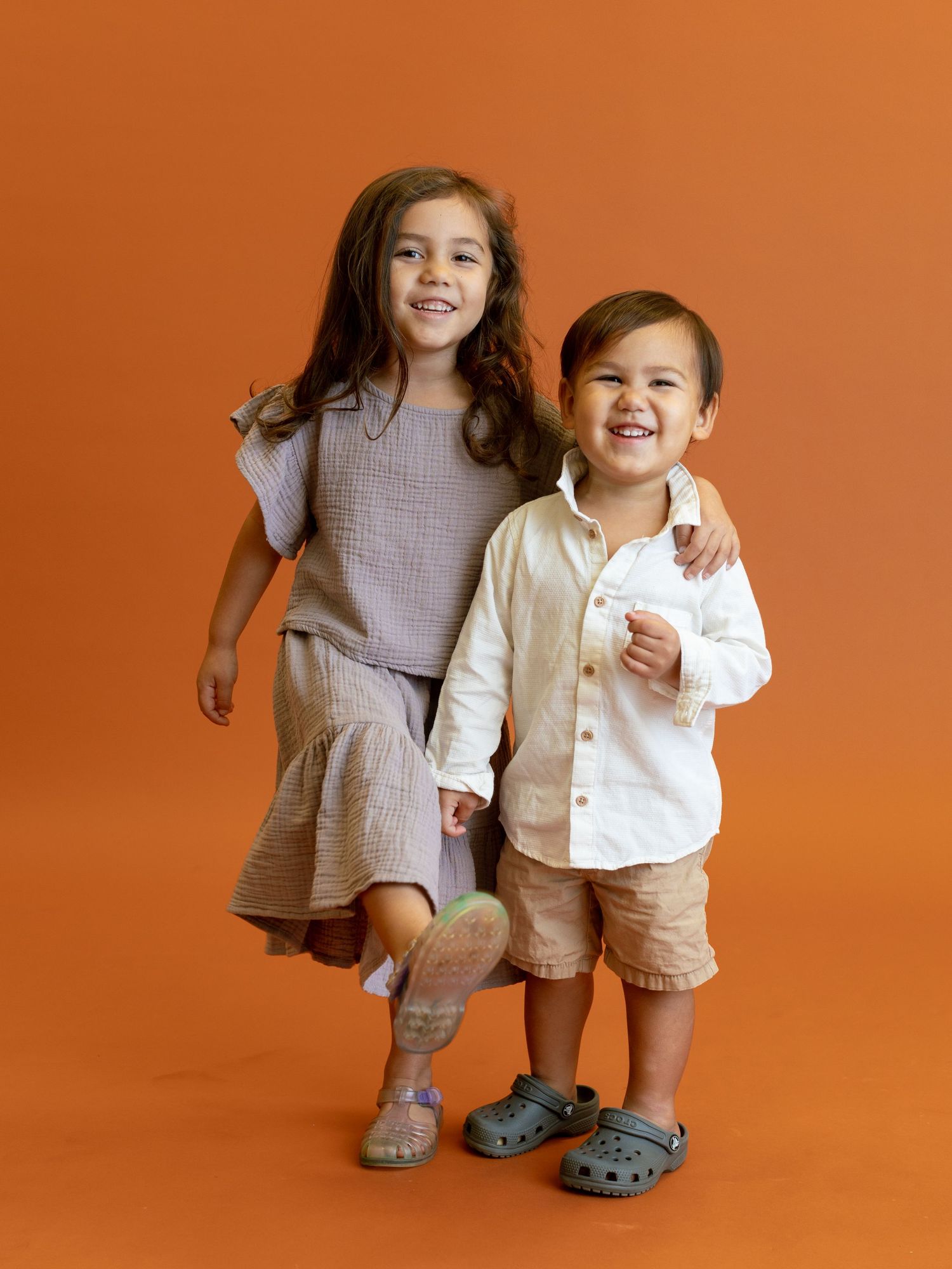 Two children in casual outfits posing together against orange background.
