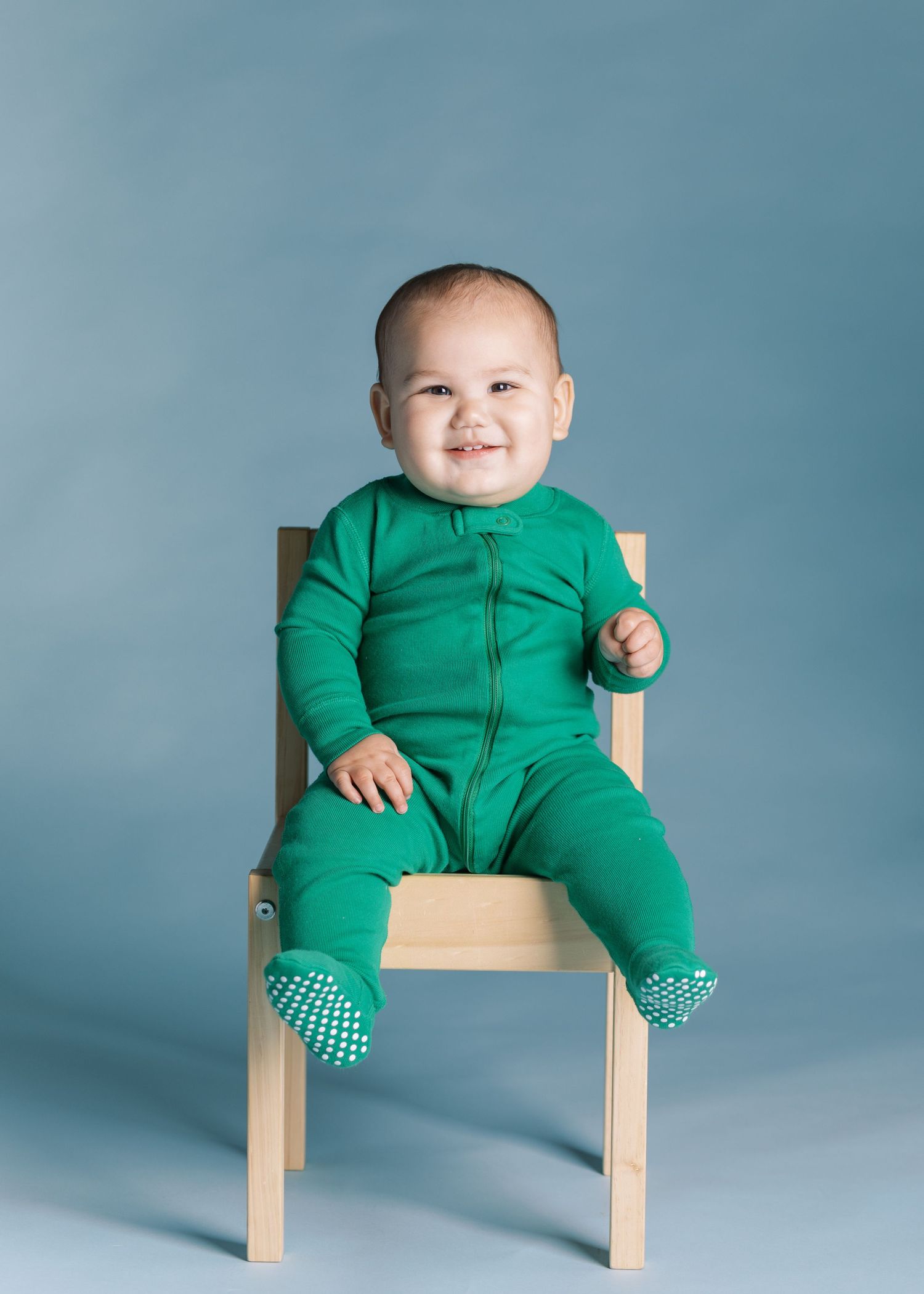 Baby wearing green onesie sitting in wooden chair against blue background.