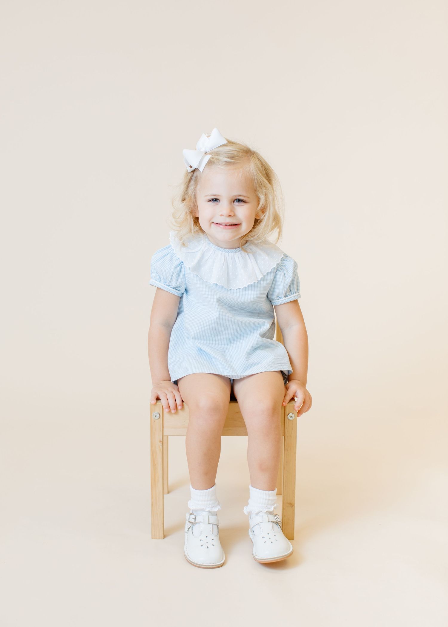 Small child in light blue dress with white bow sitting on wooden chair against beige background.