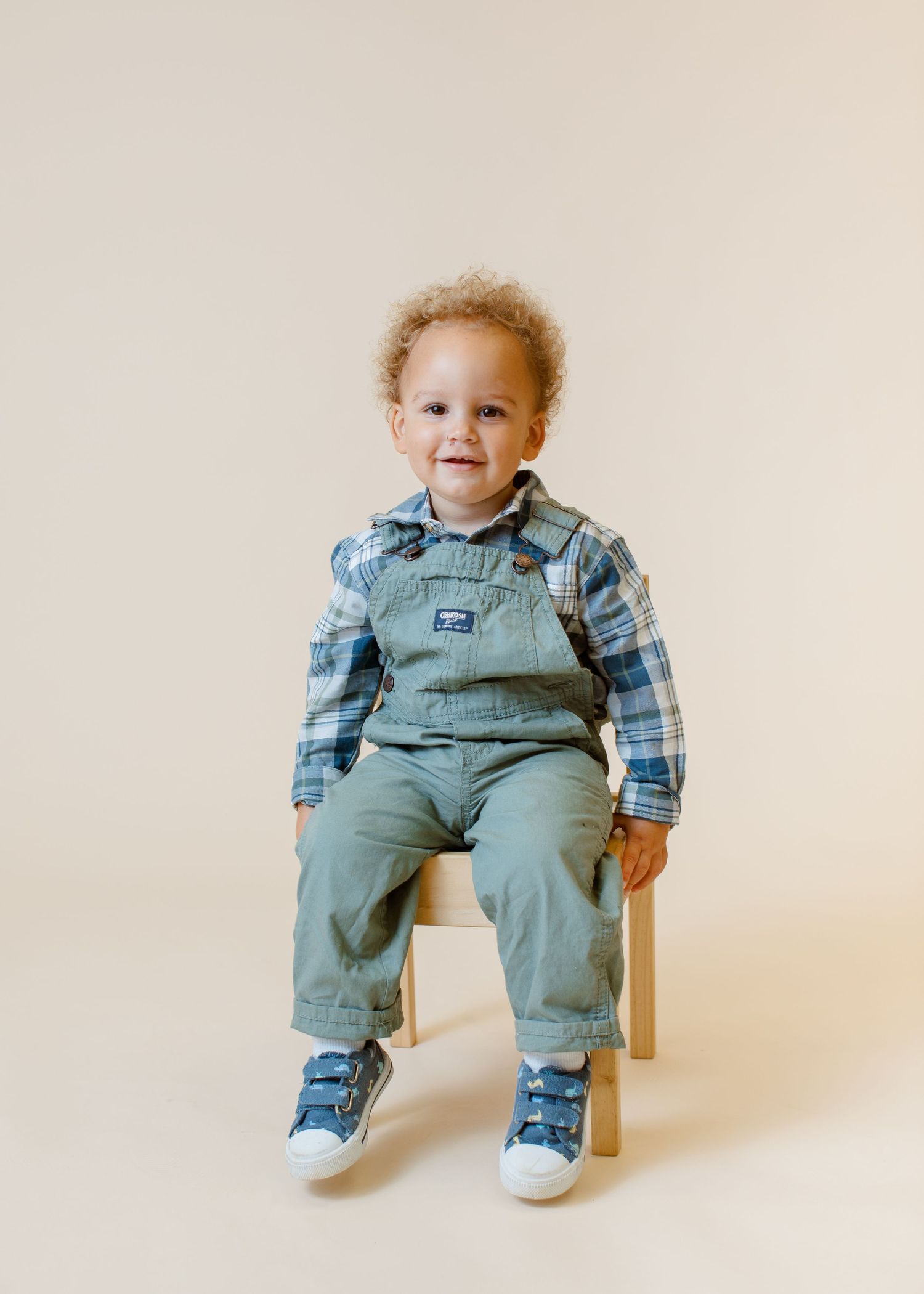 Child in plaid shirt and green overalls sitting on wooden chair with happy expression.