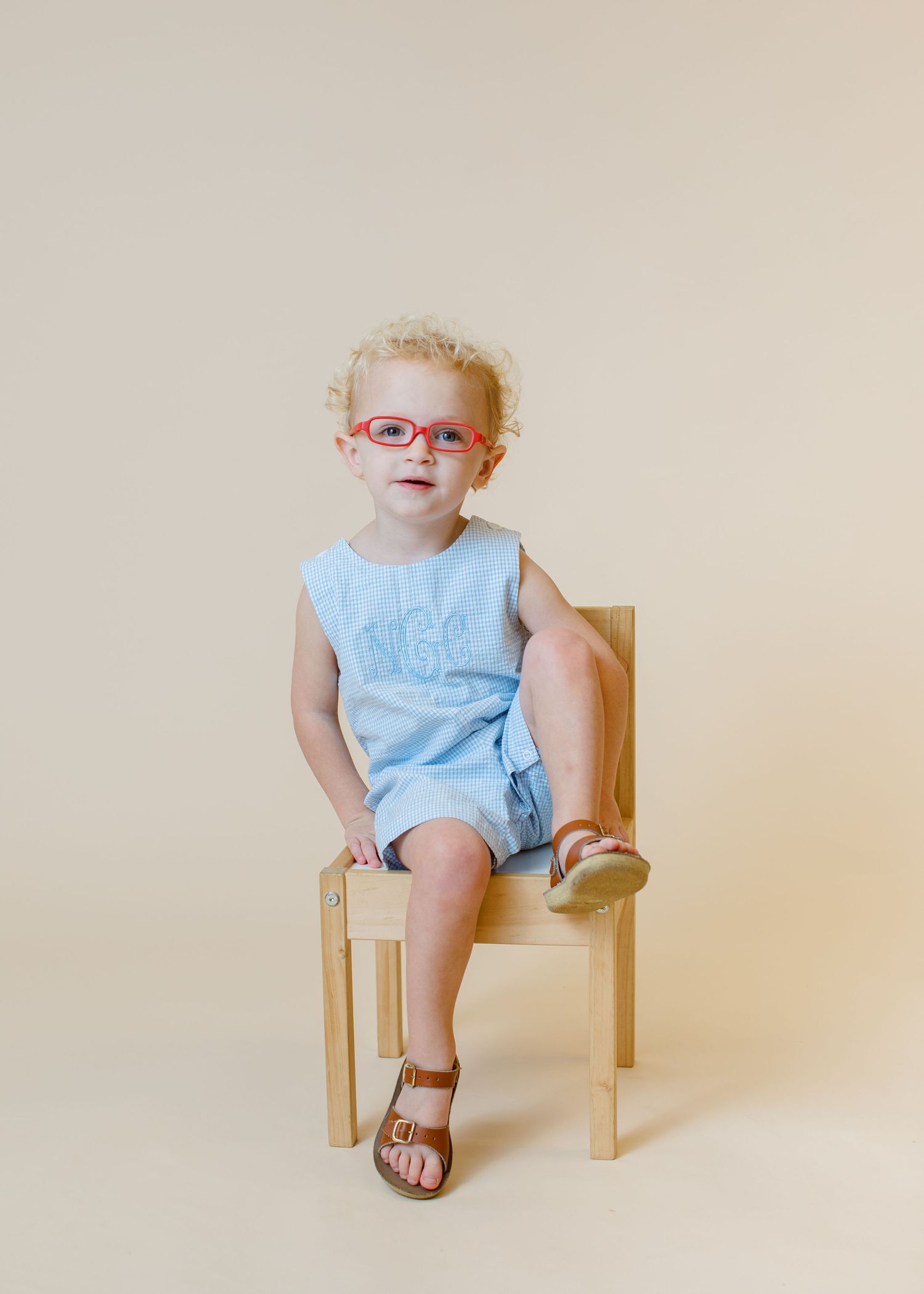 Child in light blue outfit and red glasses sits posed on wooden chair.