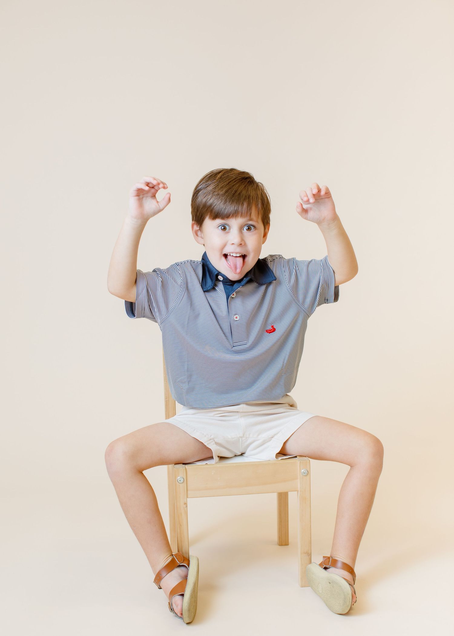 Child in striped polo making silly face while sitting on wooden chair.