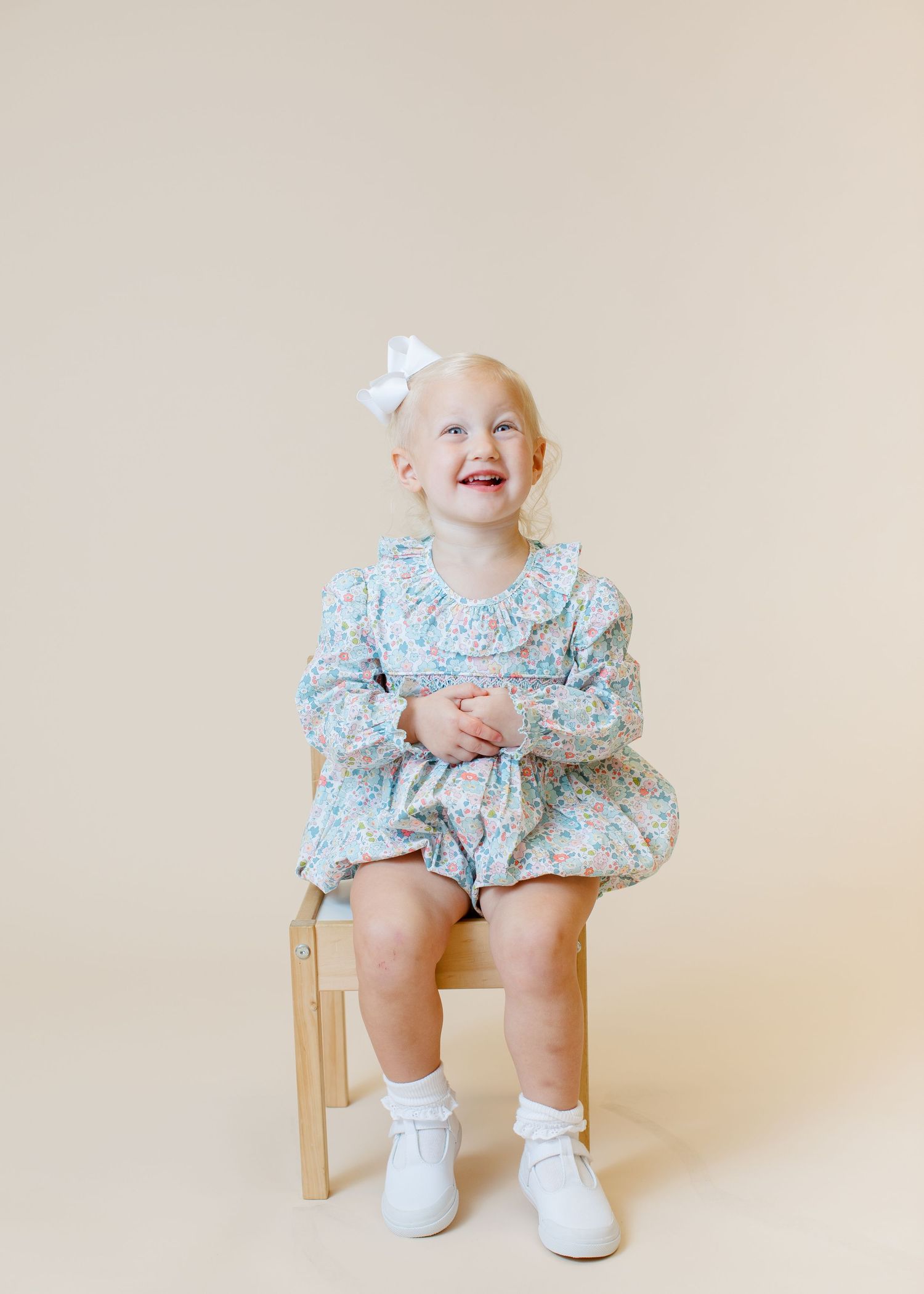 Portrait of a joyful child in floral dress and bow sitting on wooden chair against beige background.