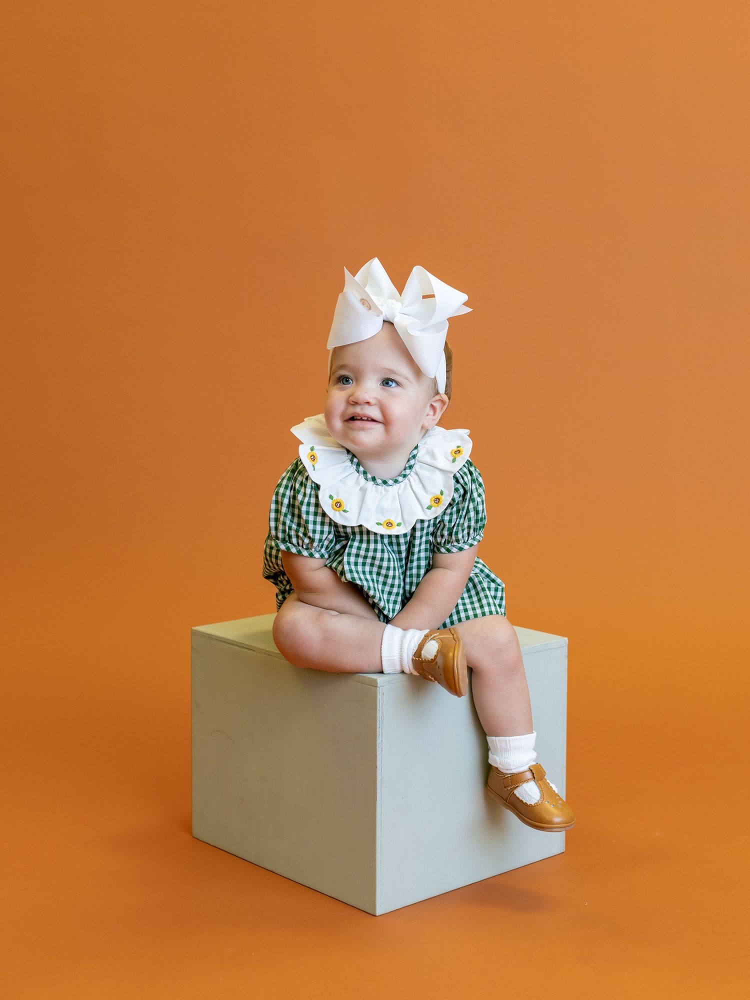 Baby in green gingham outfit with white bow sitting on cube against orange background.