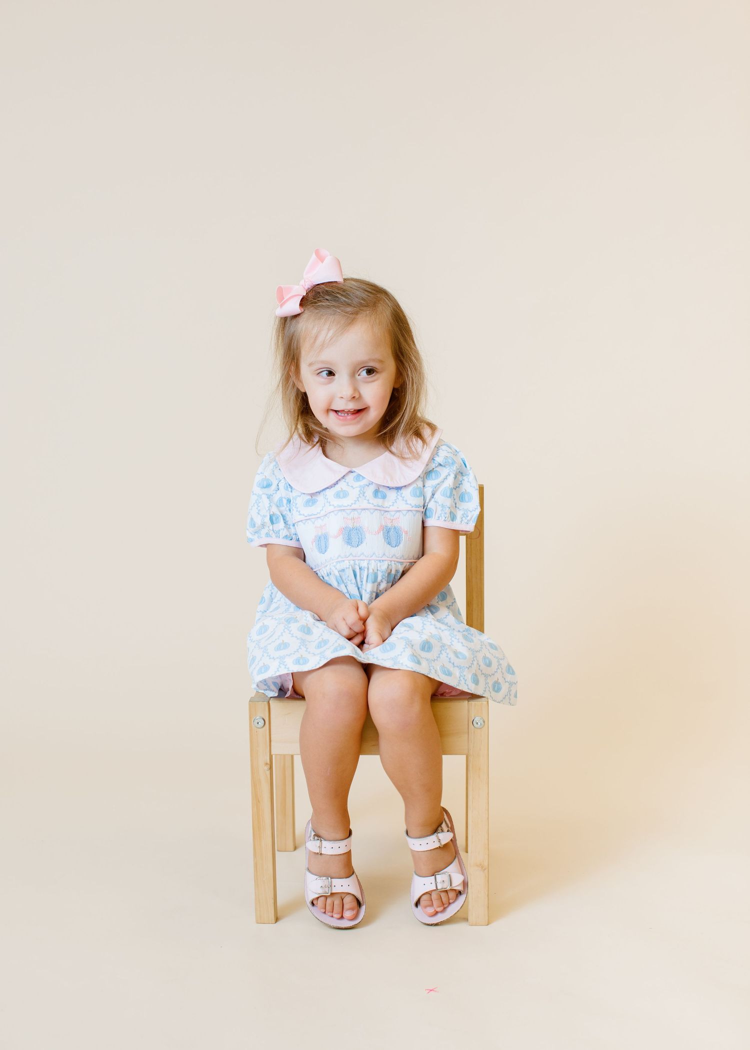 Child in blue and white dress with pink bow sits properly on wooden chair.