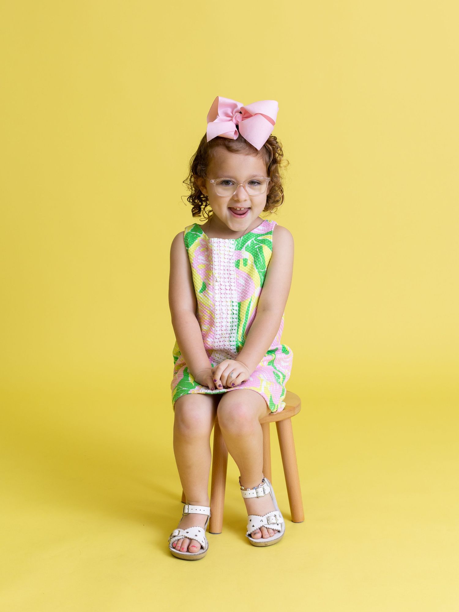 Young person in floral dress with pink bow sitting on wooden stool against yellow wall.