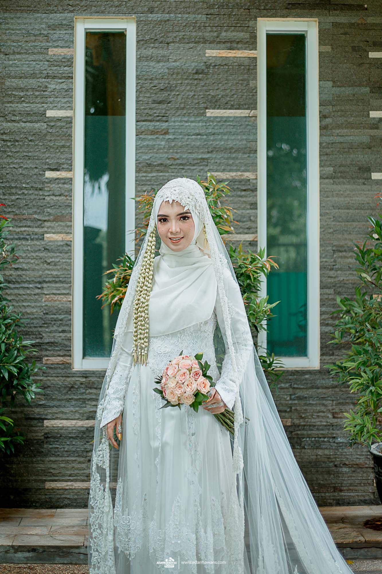 Muslim bride in an elegant white gown and hijab holds a pink bouquet against a modern architectural backdrop.