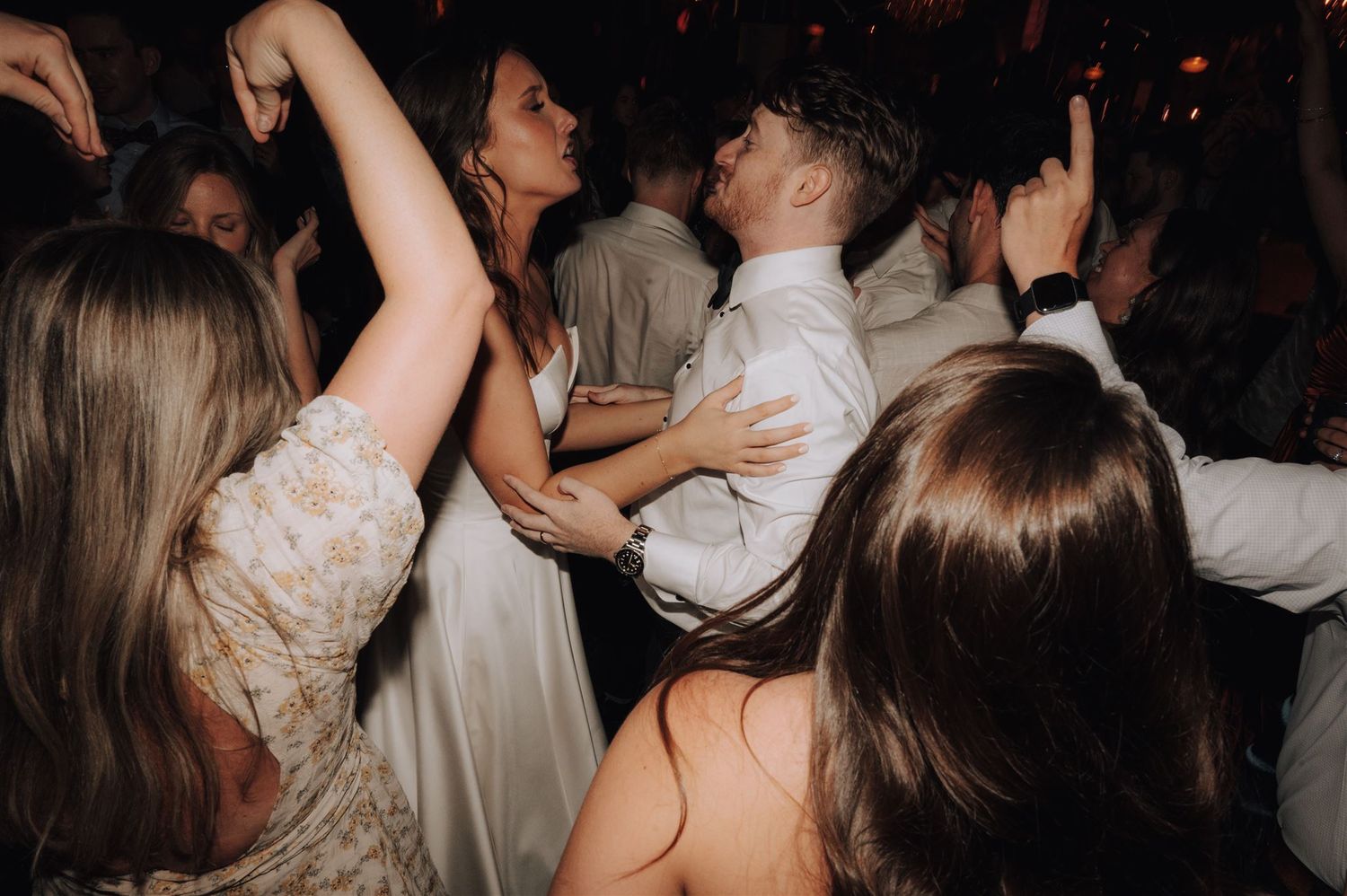 Wedding party dances joyfully under ambient lighting at reception celebration.
