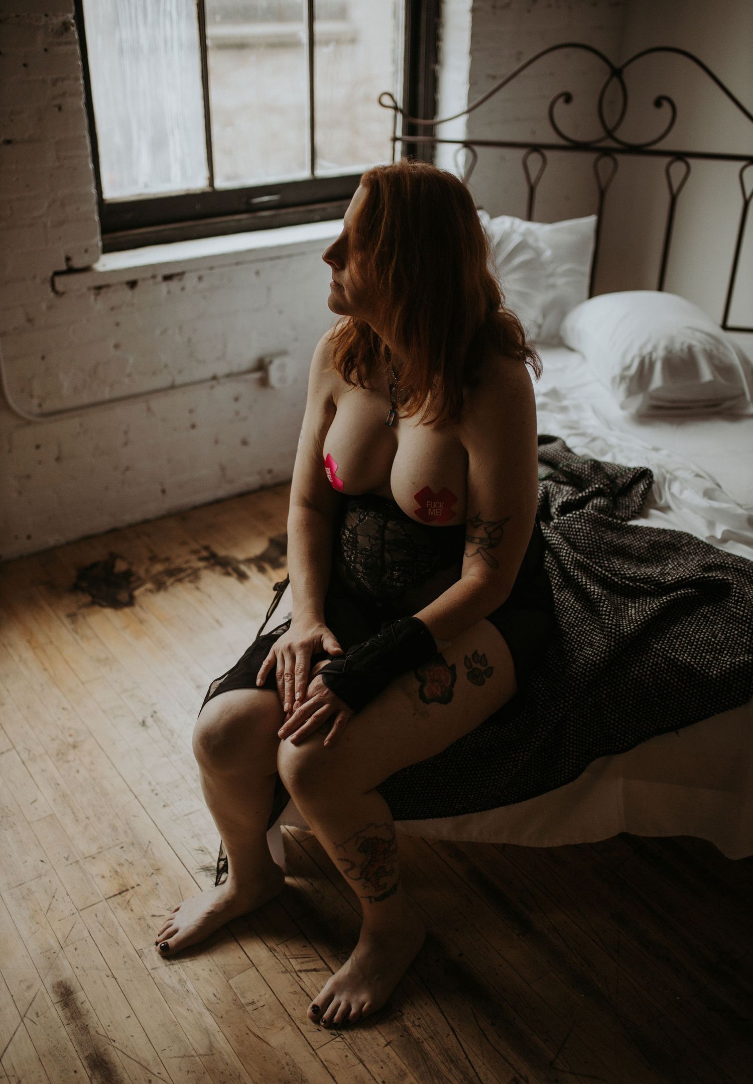 Moody boudoir photo of a trans woman in purple lingerie sitting on bed in industrial loft space with moody lighting.