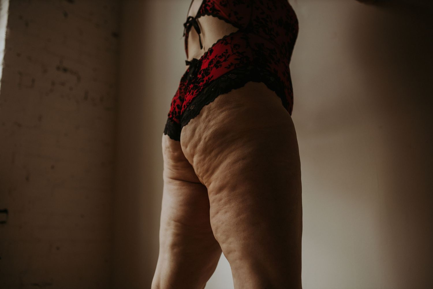 Side view of legs and torso wearing red lingerie against a dark wall in moody lighting.