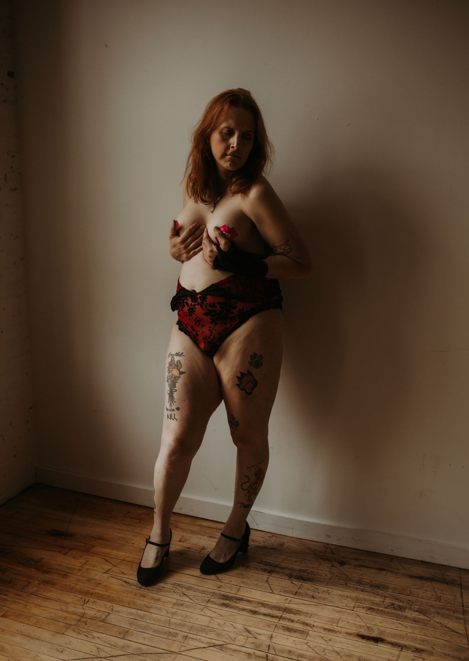 Artistic boudoir photo featuring dark lingerie and heels on wooden floor with moody lighting against neutral wall.