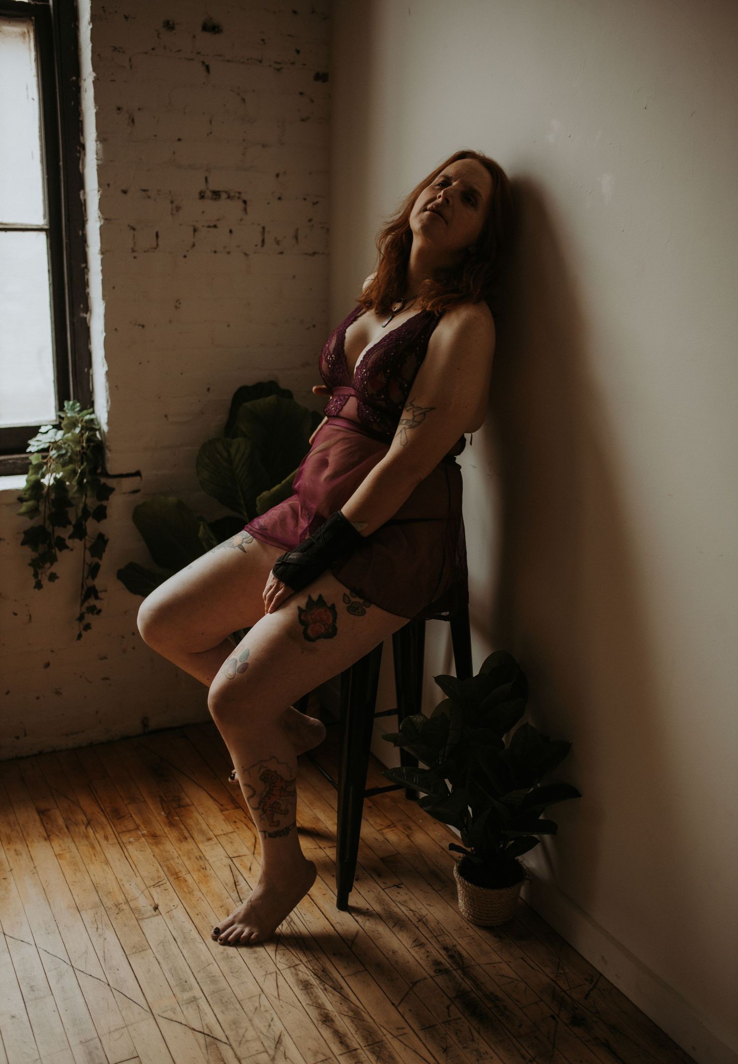 A transfemme person wearing a purple slip dress poses against a white wall near a window and house plants.