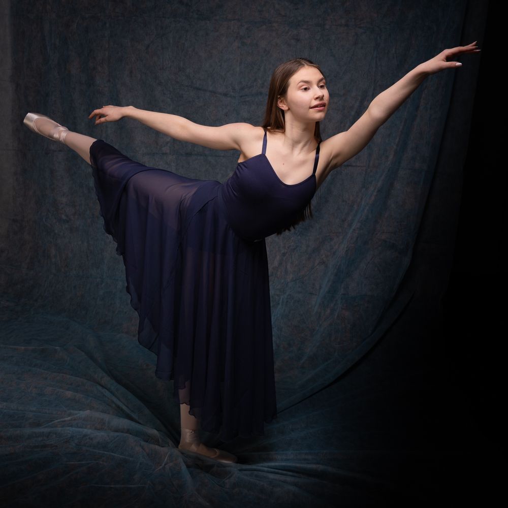 Ballet dancer in navy dress performing graceful arabesque pose against dark backdrop.