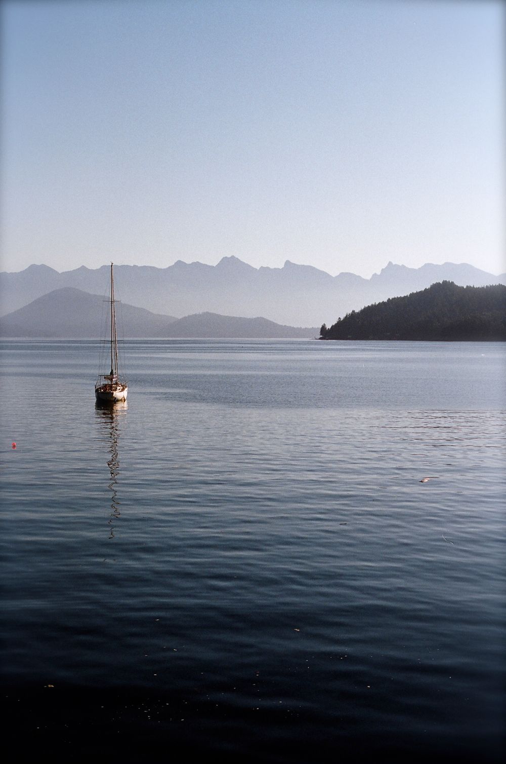 Sailboat anchored on calm water with misty mountain silhouettes in the background in Robert's Creek, BC Canada, 35mm film.