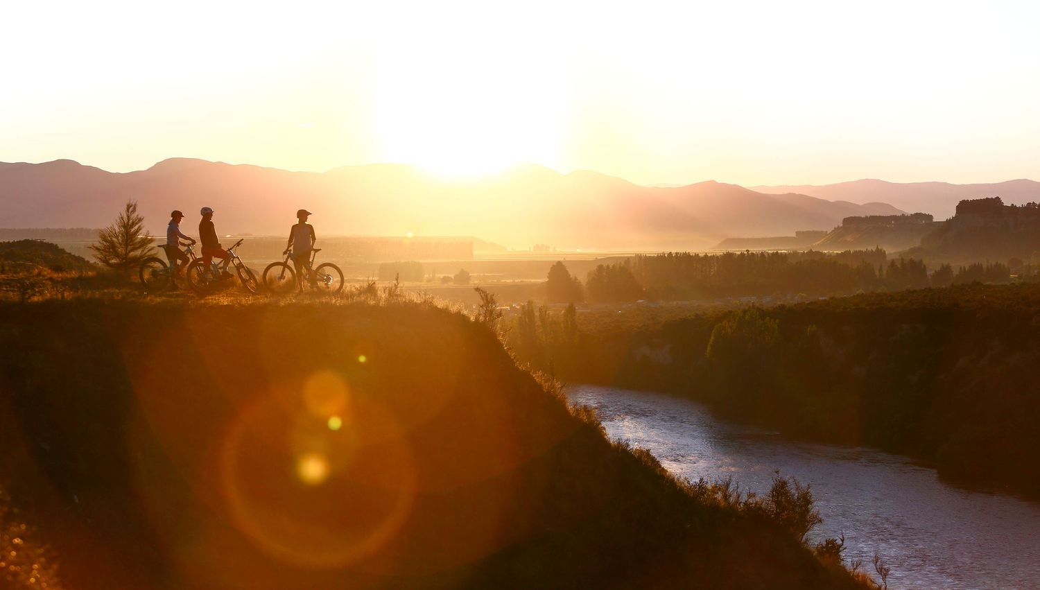 Cyclists silhouetted against a golden sunset overlooking a scenic river valley with misty mountains.