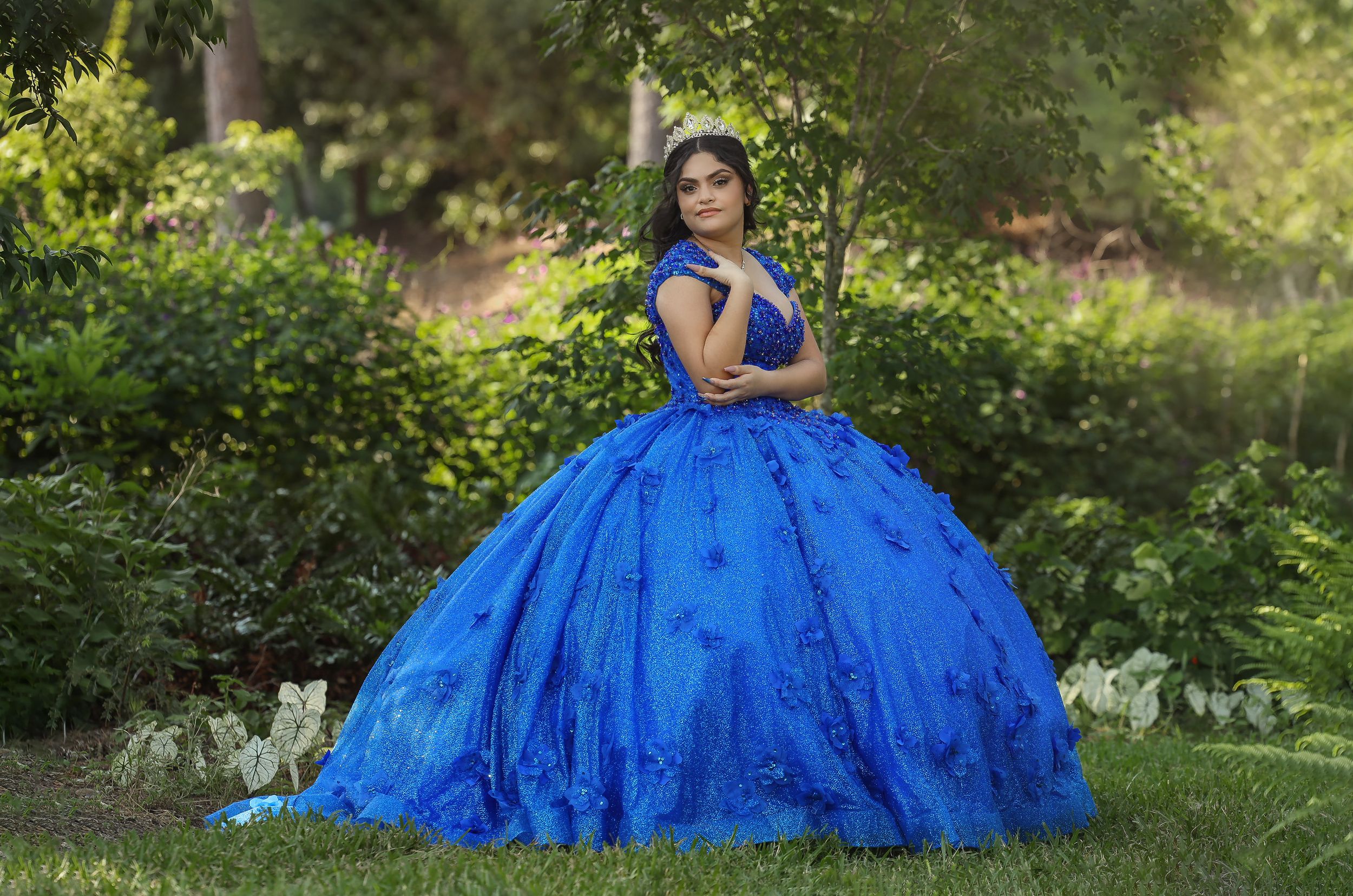 A stunning royal blue quinceañera gown photographed in a lush garden setting.
