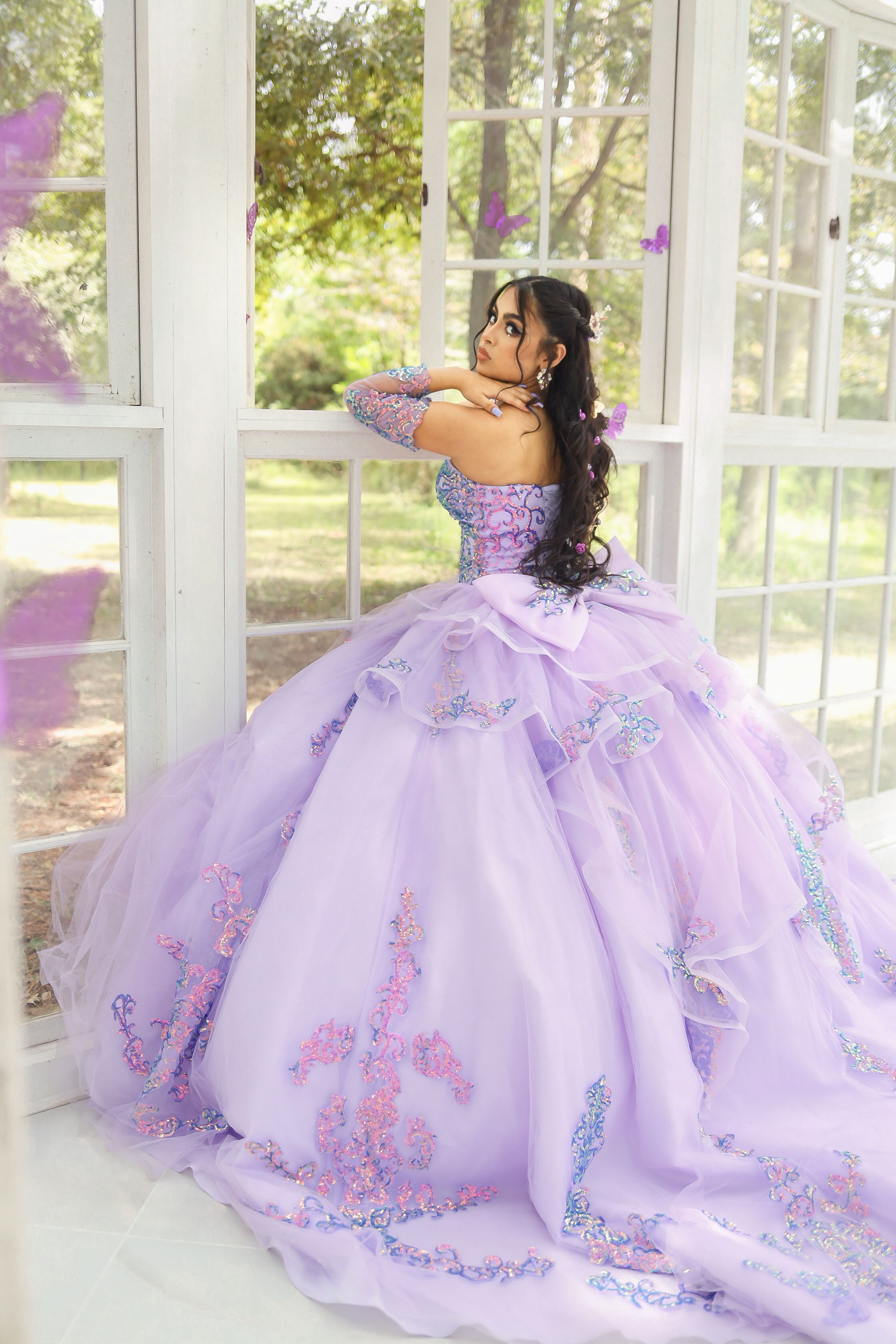 A lavender quinceañera ball gown with butterfly details sparkles in a sunlit conservatory.