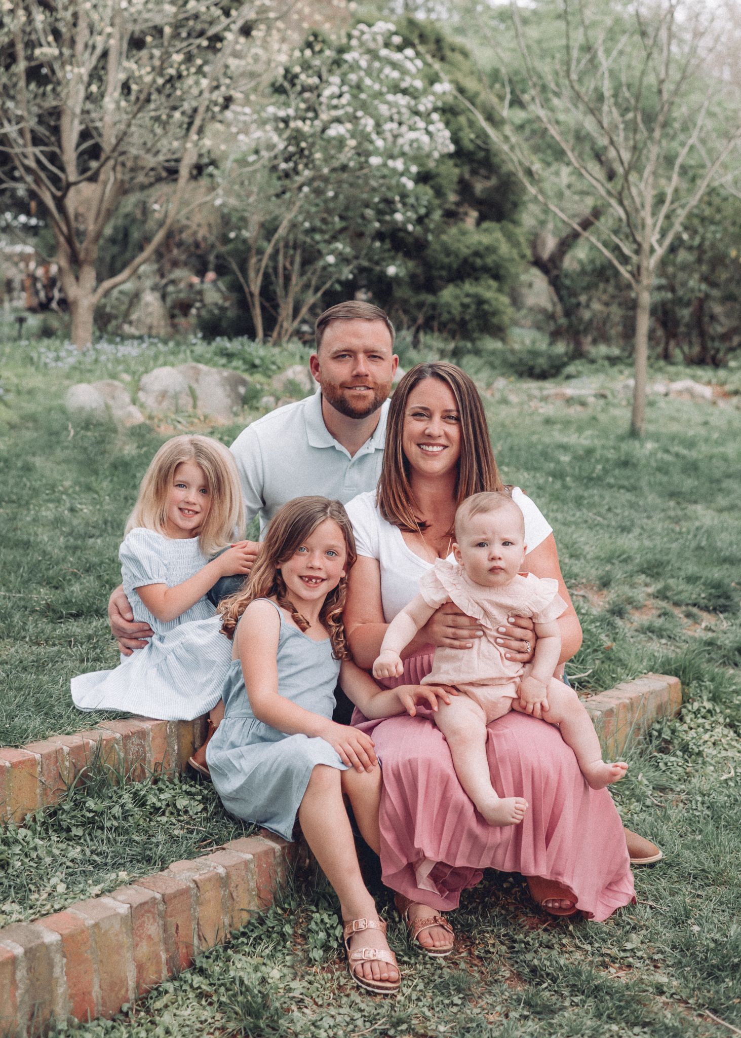 A family of five sits together on a brick ledge in a garden setting.