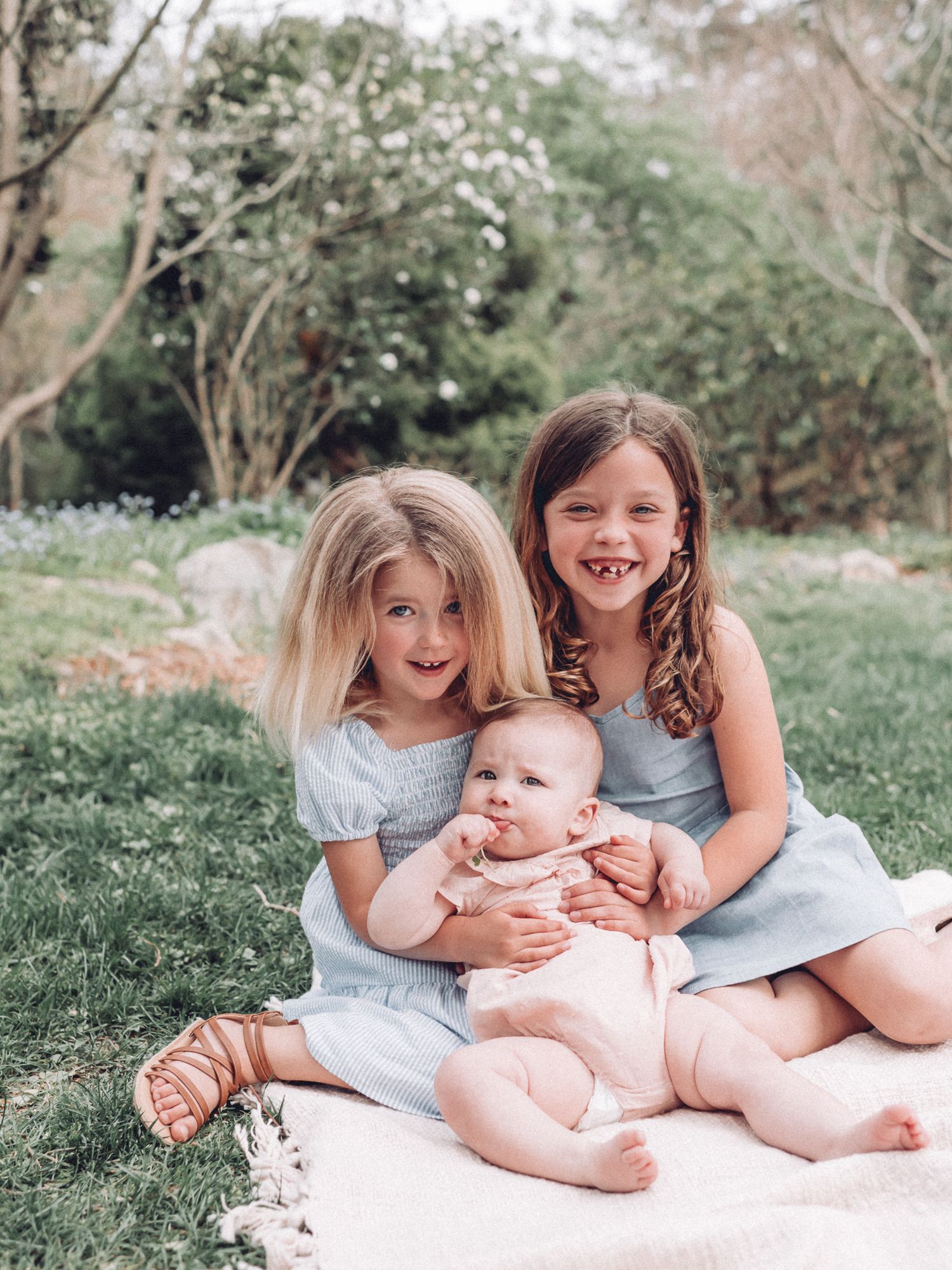 Three siblings in soft blue outfits share joyful moments on a blanket outdoors.