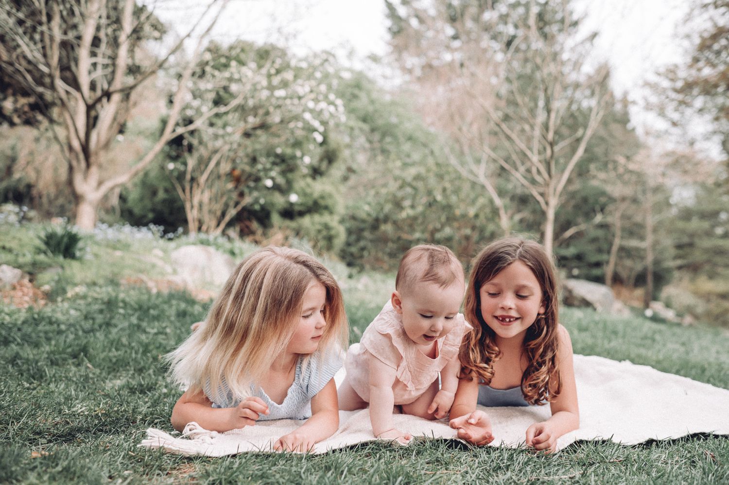 Three children lying on their stomachs on a white blanket in a lush garden setting.