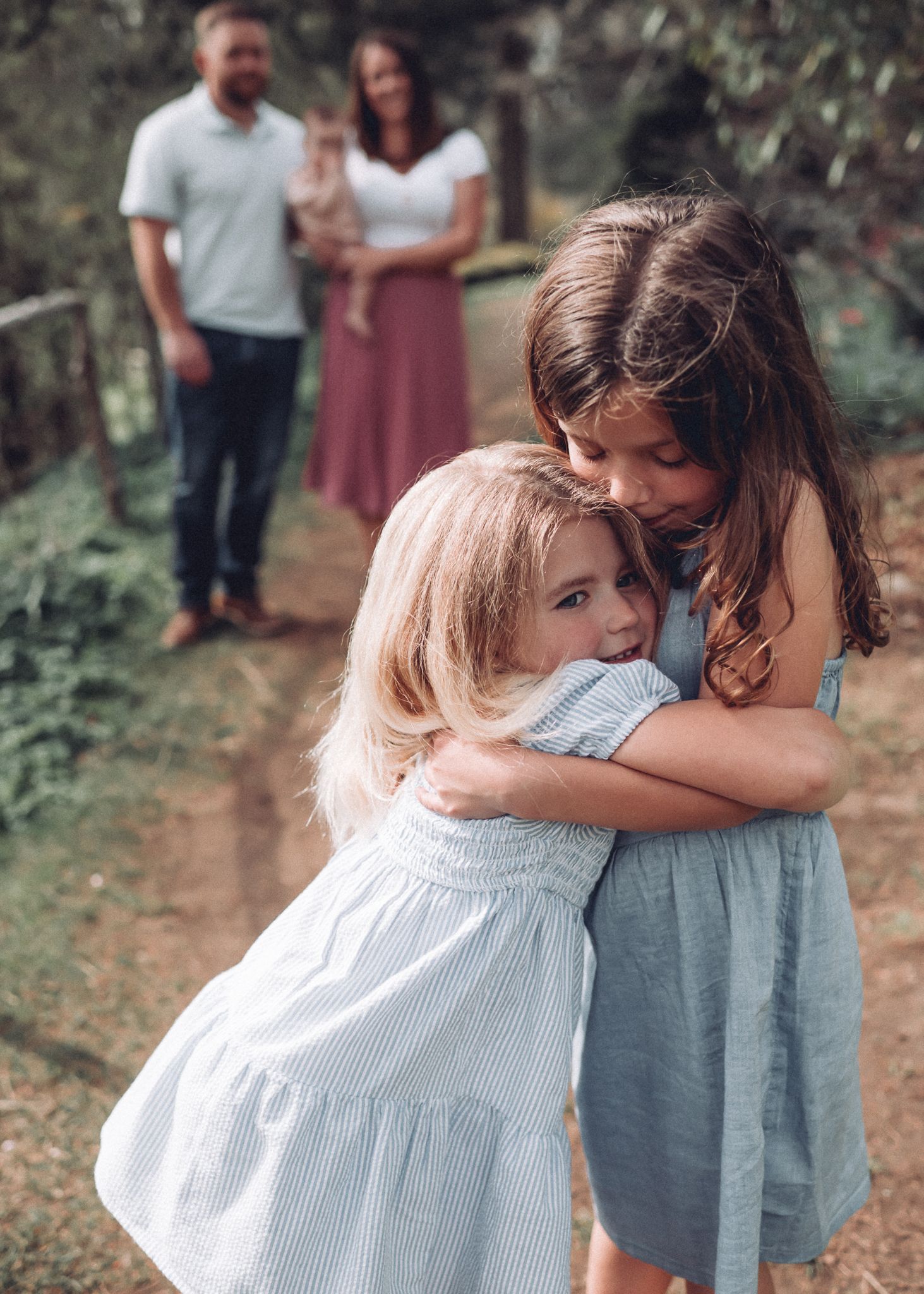 Siblings share a loving embrace while adults look on in garden background.