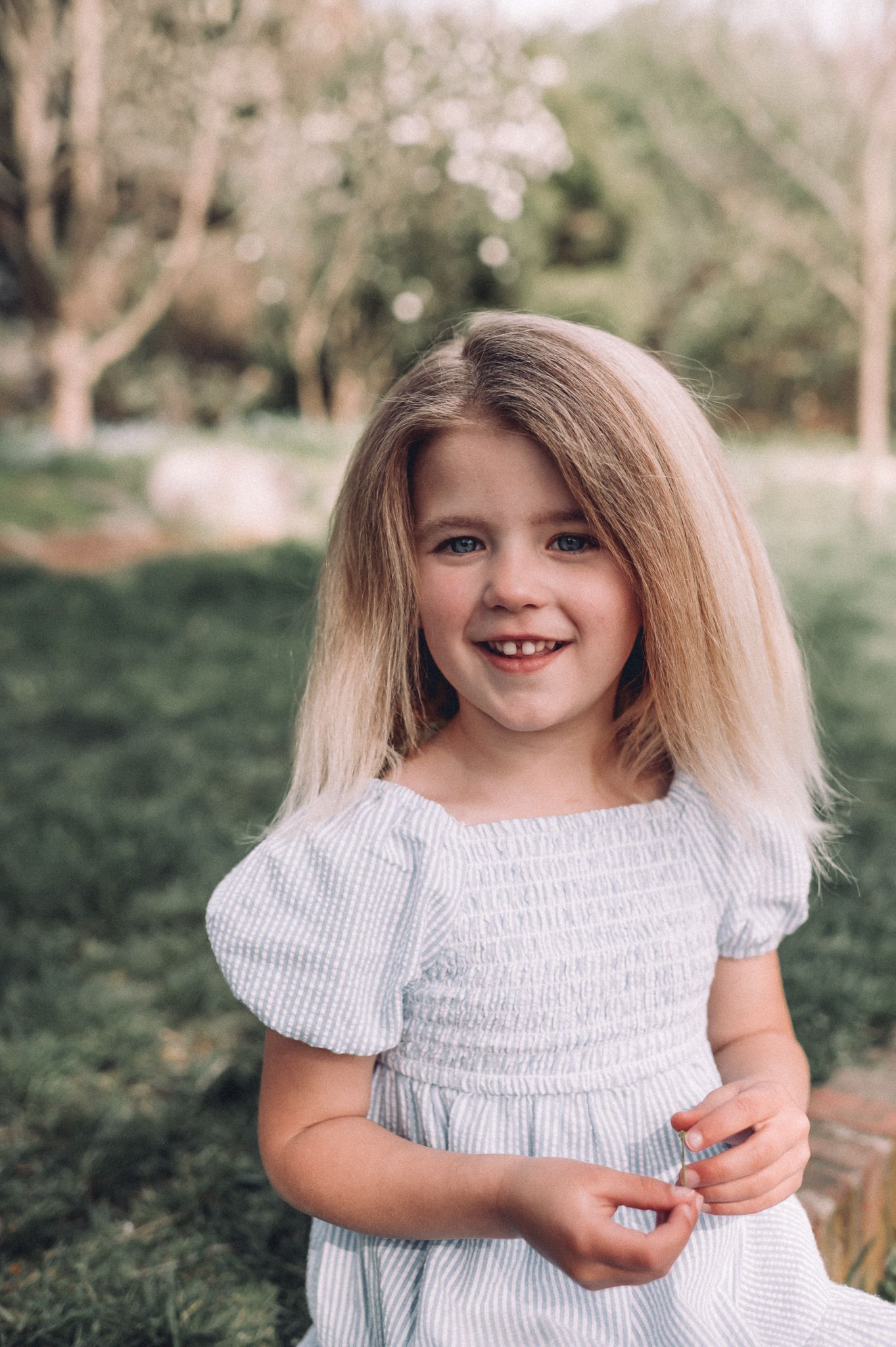 Young child in a white smocked dress smiles during an outdoor portrait session.