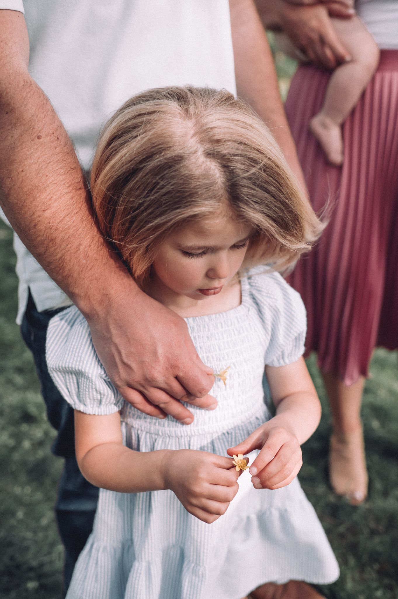 Child in white dress looking down while holding something small in hands during outdoor photoshoot.