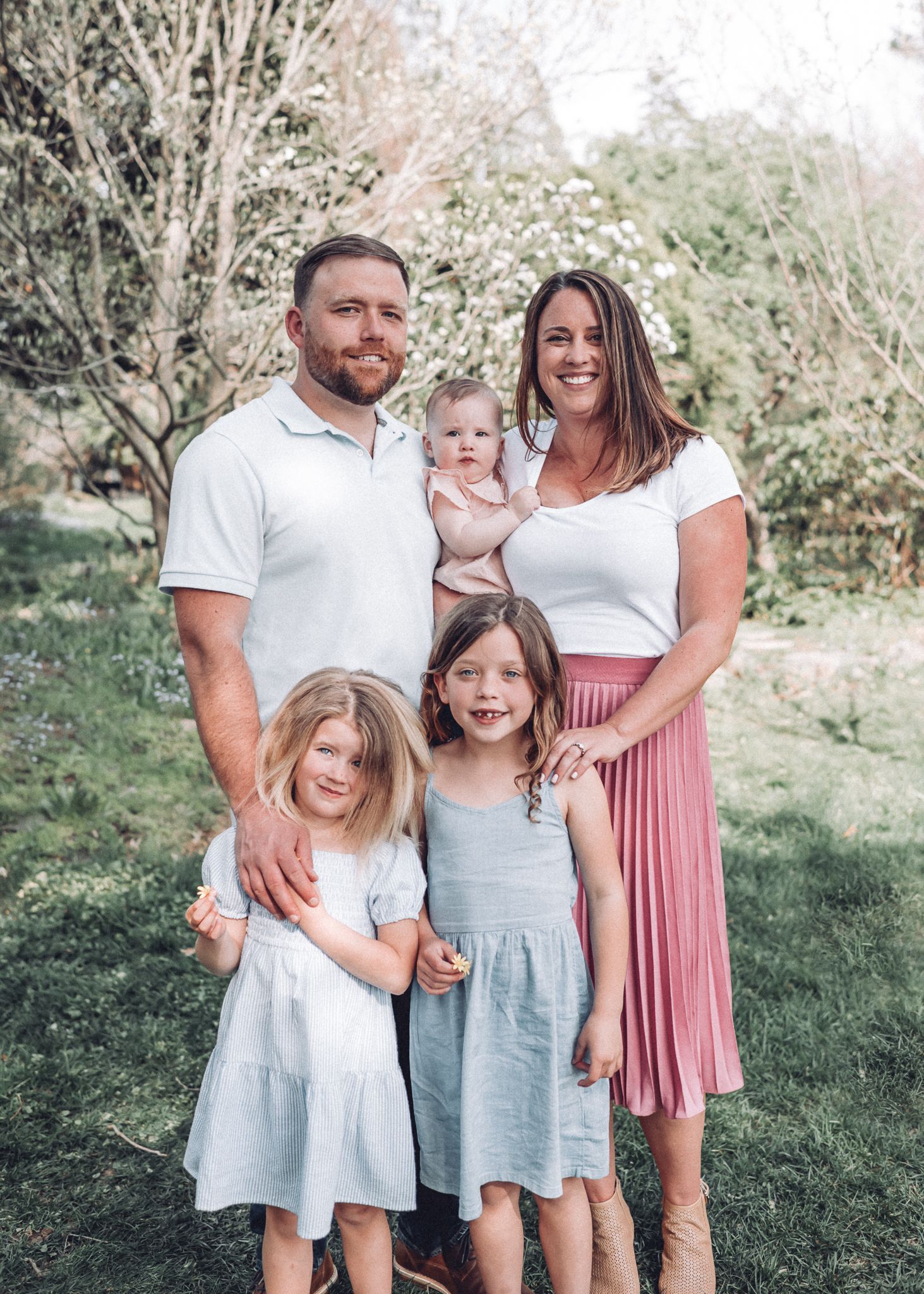 Family wearing coordinating outfits poses together in a grassy outdoor setting on a sunny day.
