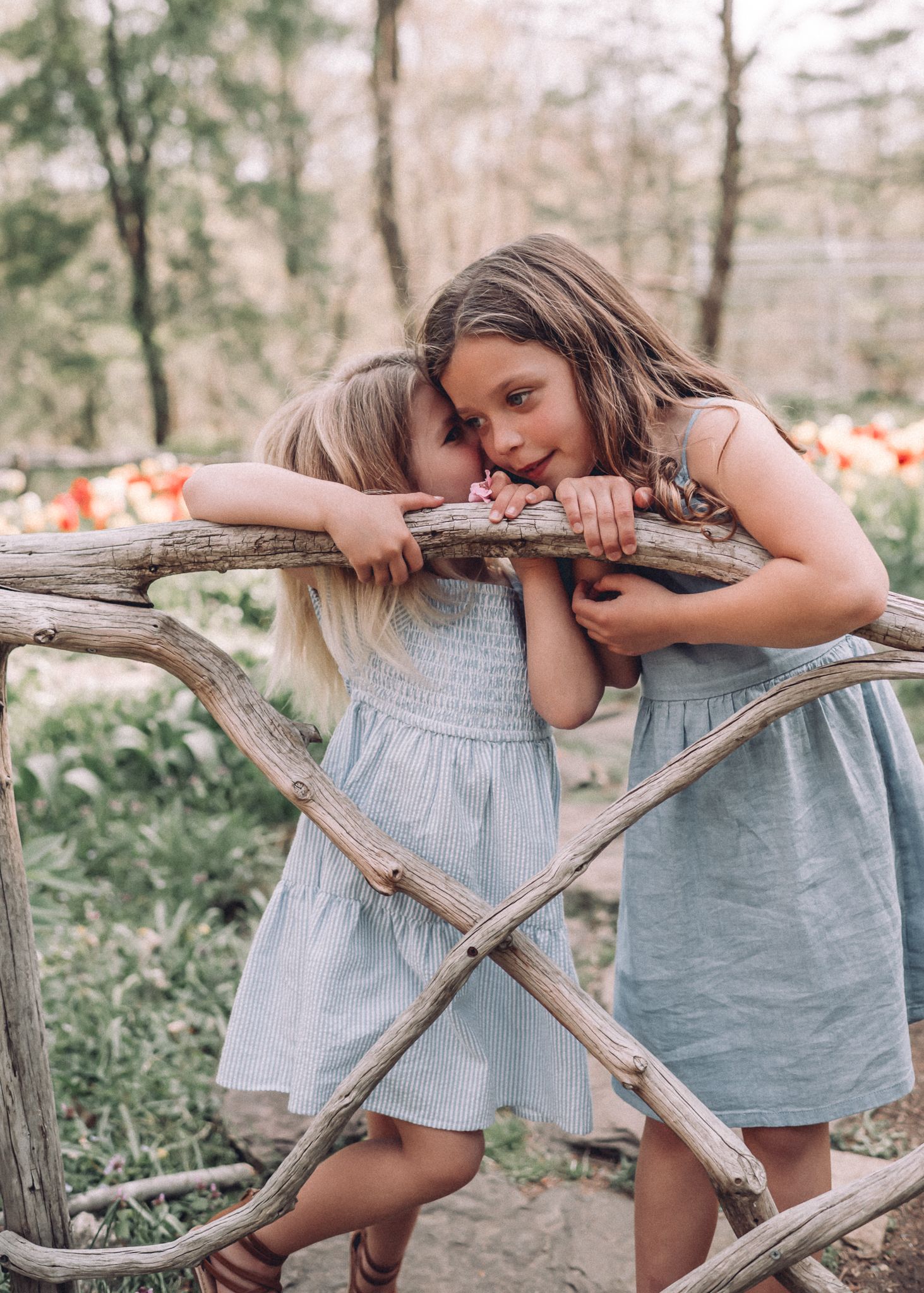 Two children in matching dresses playfully lean on rustic wooden fence in garden setting.