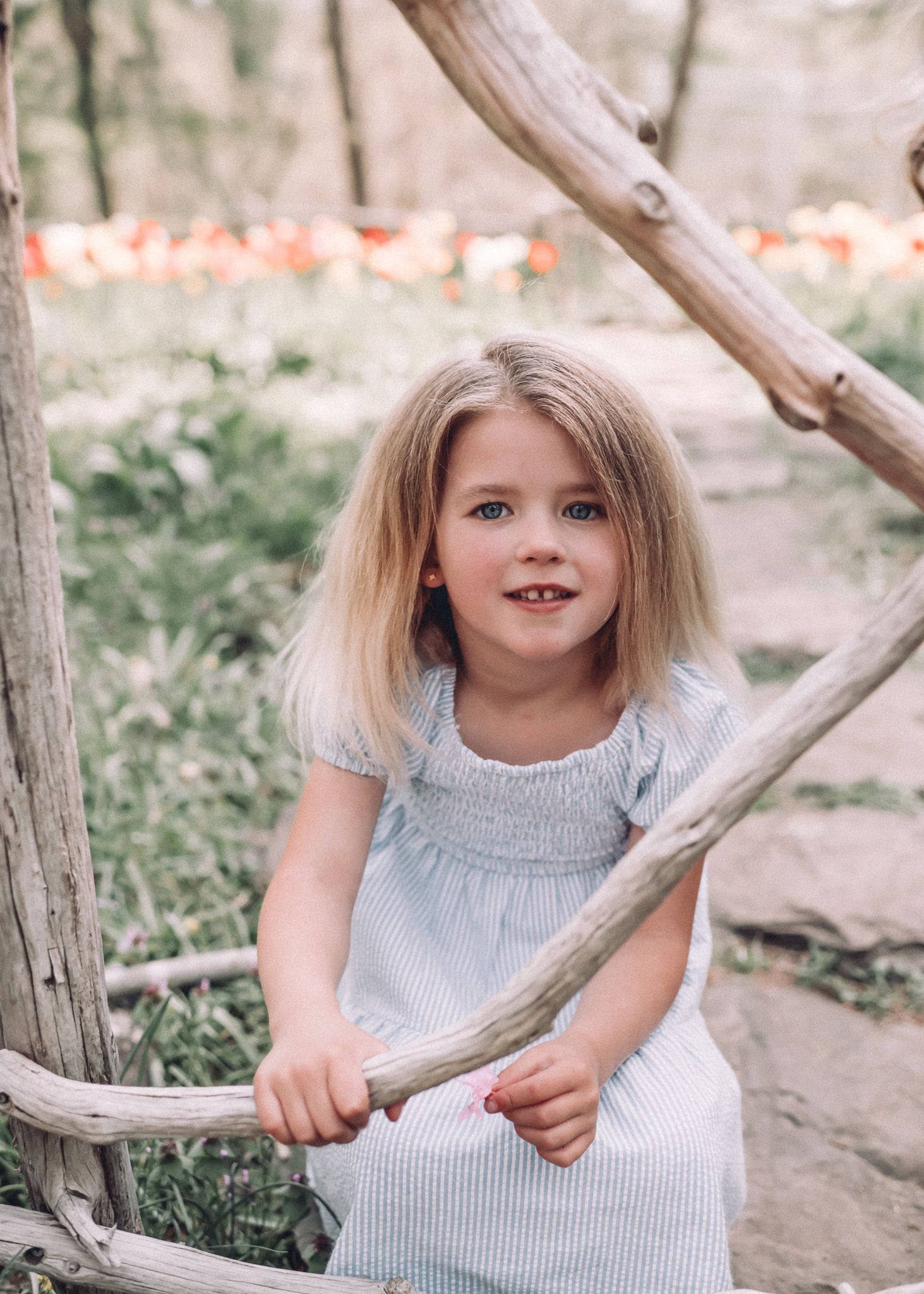 Child in striped dress smiles while playing on wooden garden fence structure.