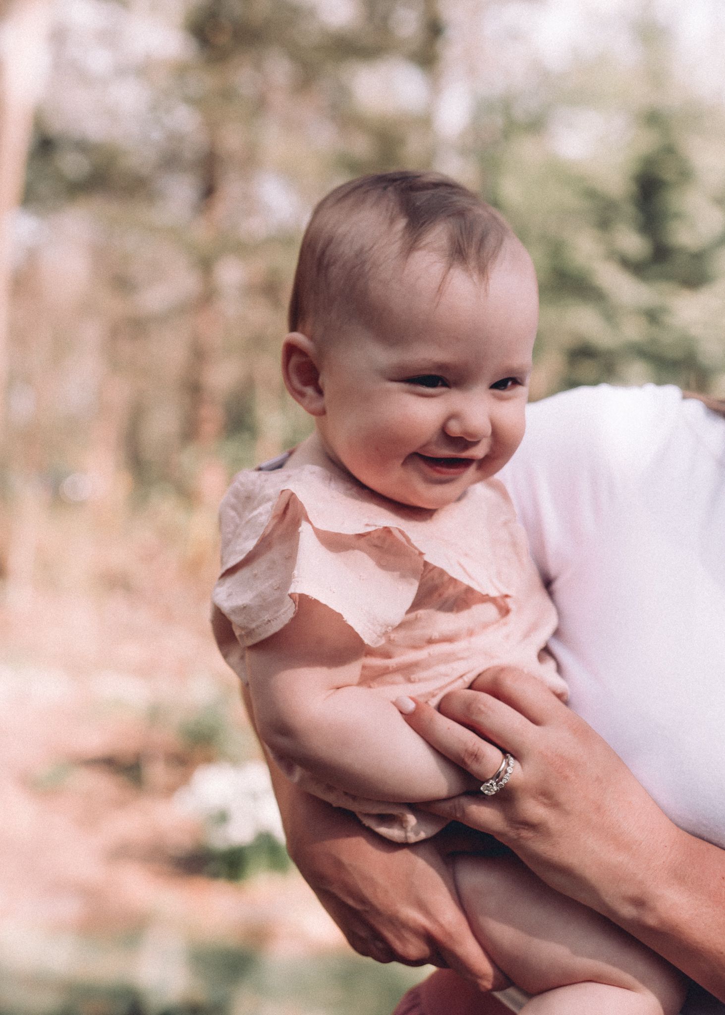 A baby in a light pink outfit smiles joyfully while being held up by adult hands.